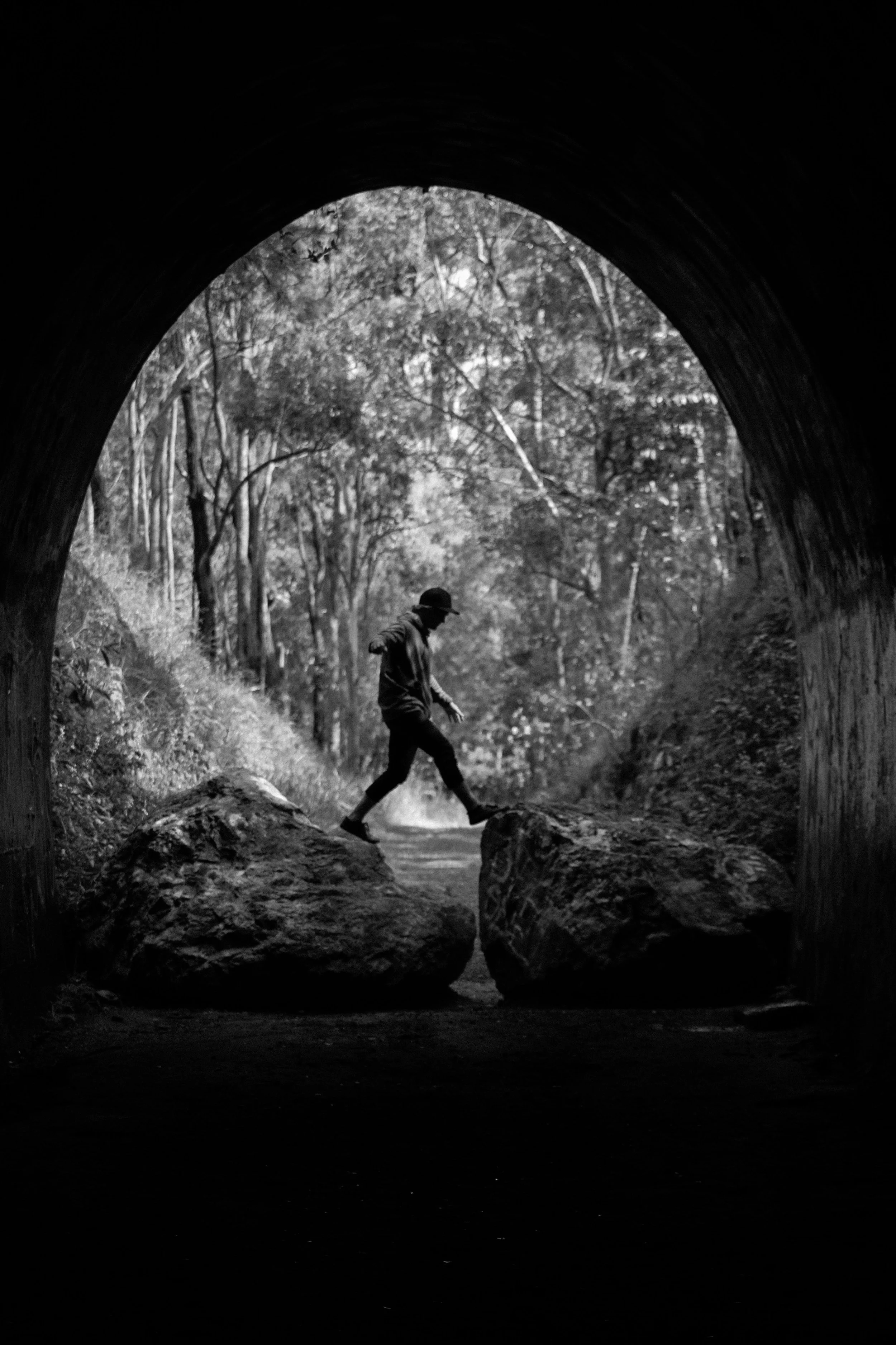A person wearing a hat and dark clothing balancing on large rocks inside a tunnel, with a wooded area visible outside the tunnel.