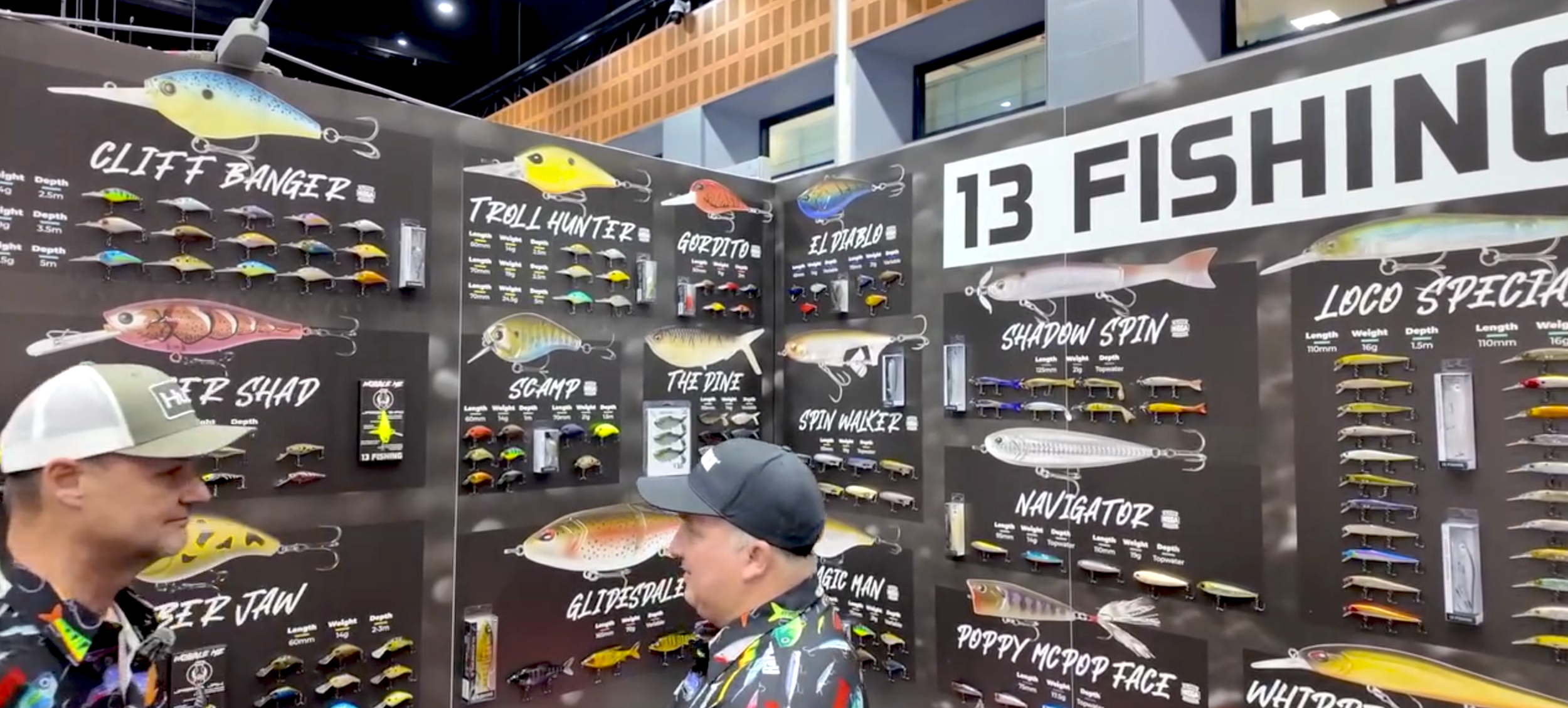 Display of various fishing lures and tackle with labels and prices, two men looking at the display, at a fishing expo or store.