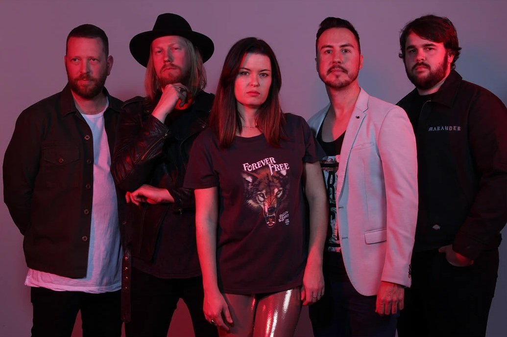 Group of five young adults standing together against a gradient background, dressed in casual and stylish clothing, with serious expressions.