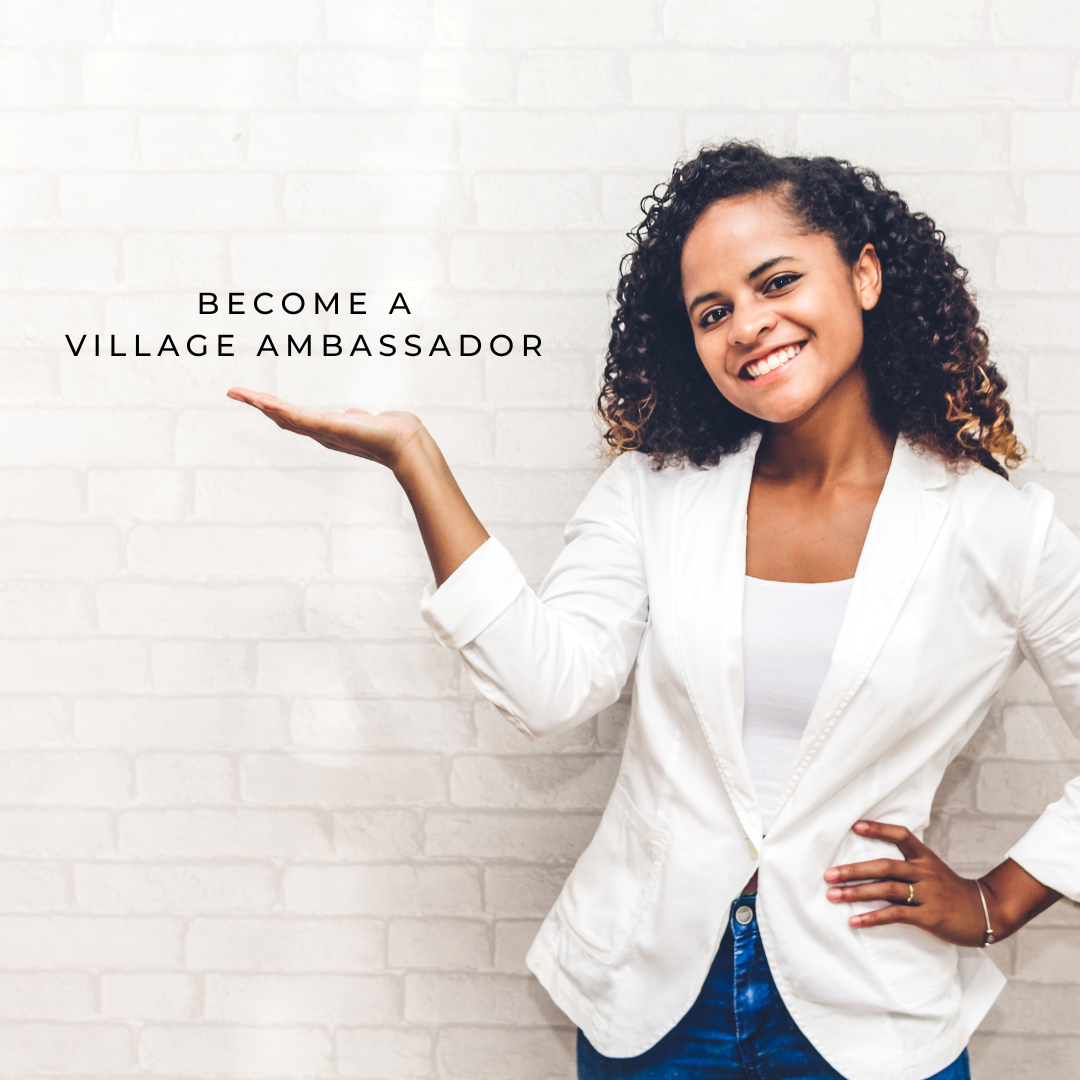 A young woman with curly hair smiling and wearing a white blazer, standing against a white brick wall, with her hand extended to the side and a text overlay that says "Become a Village Ambassador."