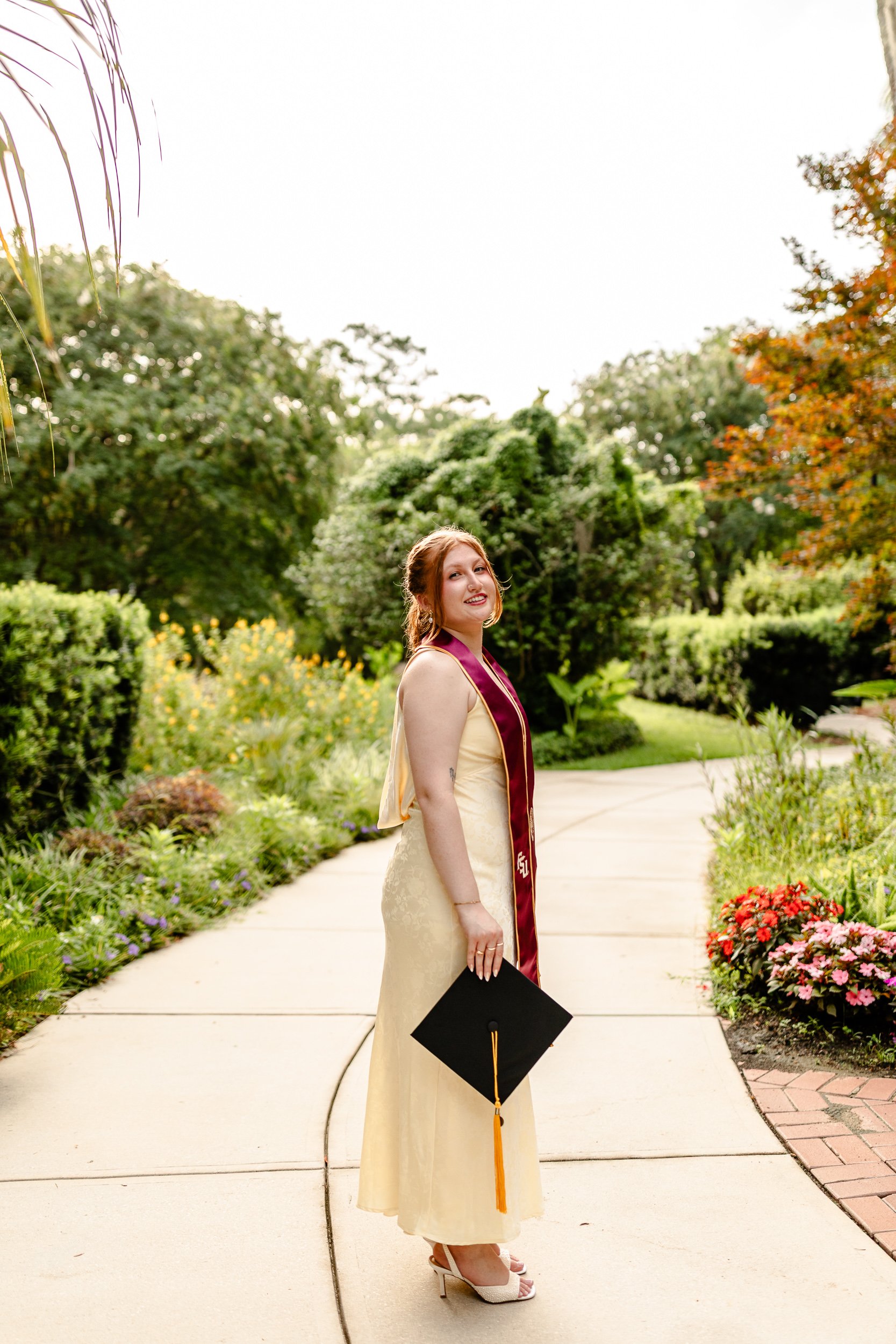 A young woman in a cream-colored dress and white high heels standing on a garden path, holding a black graduation cap with an orange tassel, smiling with trees and colorful flowers in the background.