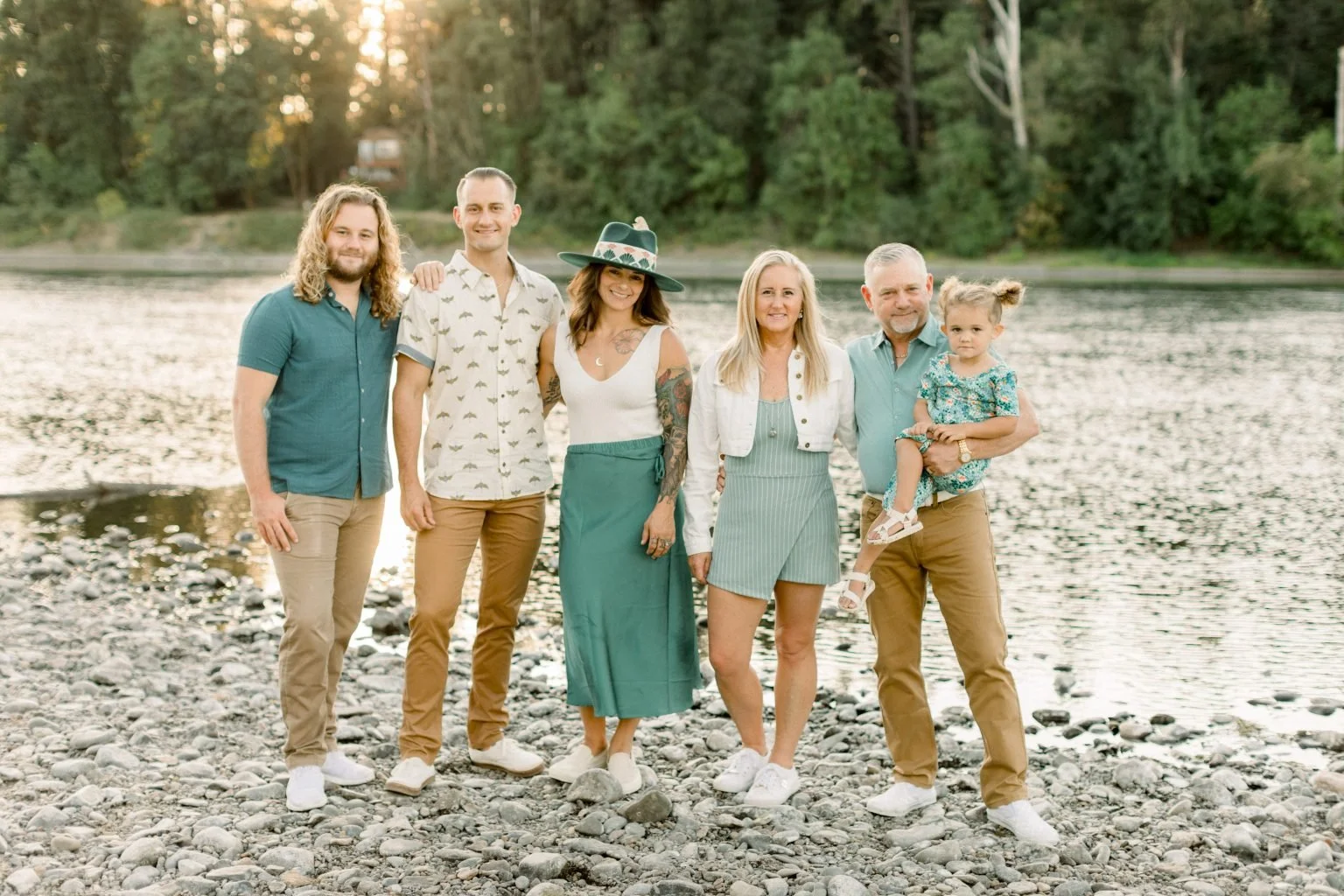 A family photo of the Thomas family of Crown Creations Dental Laboratory in McMinnville, Oregon standing on rocky riverside with trees in the background, smiling at the camera, during sunset.