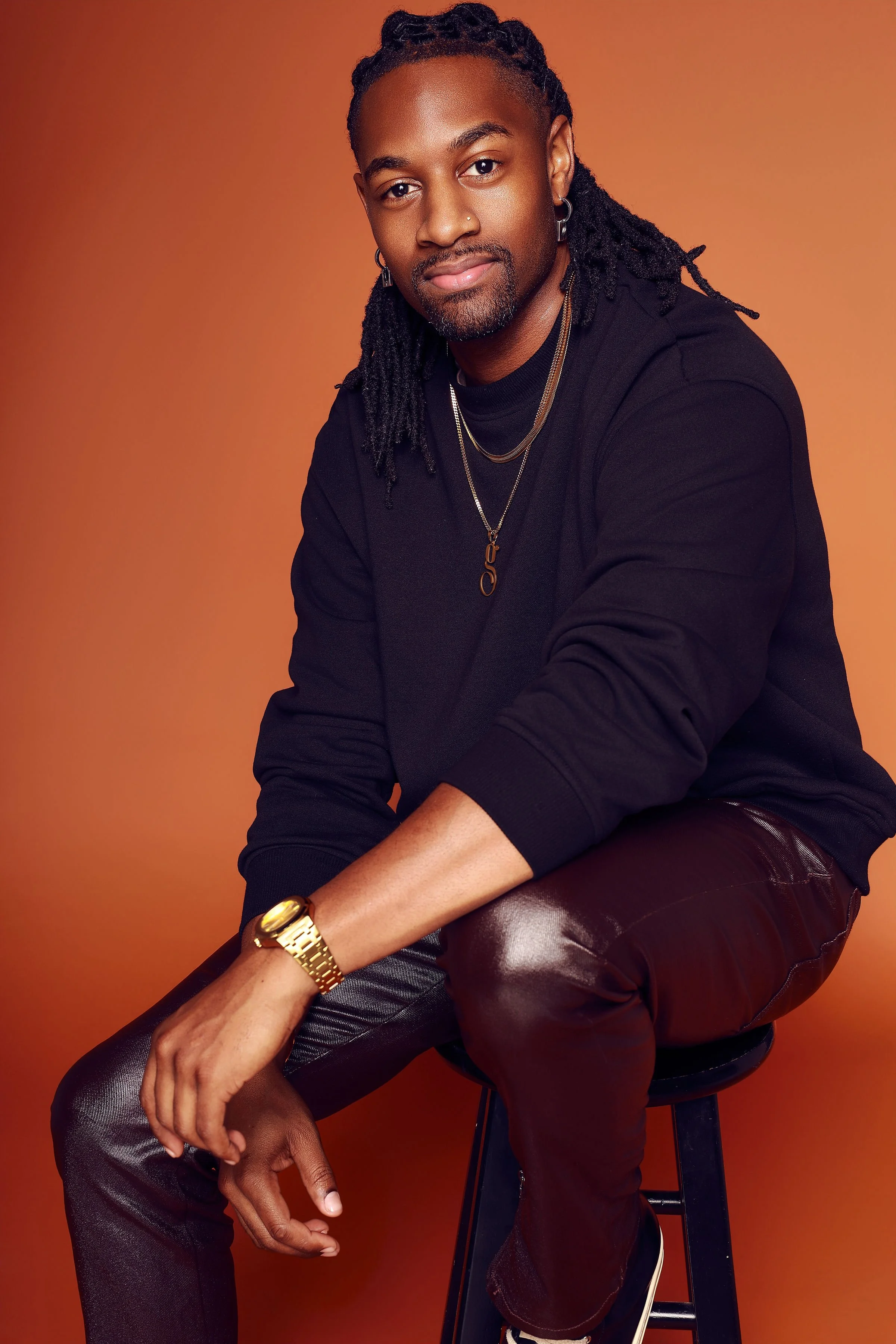 A young man with locs, beard, and mustache wearing a black shirt, gold chain, wristwatch, holding a phone, standing against a plain white background.