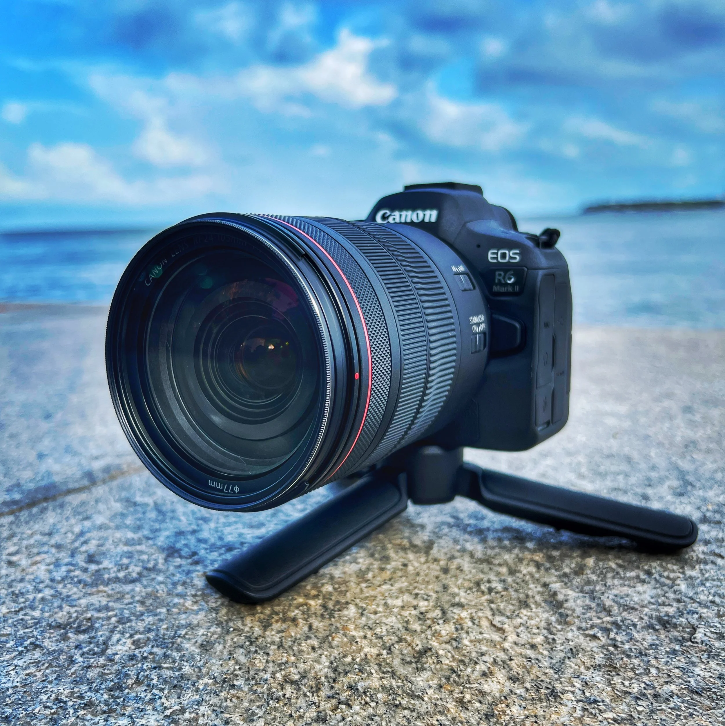 A Canon EOS R6 Mark II camera with a large telephoto lens mounted on a small tripod, placed on a concrete surface near a body of water with a cloudy sky.