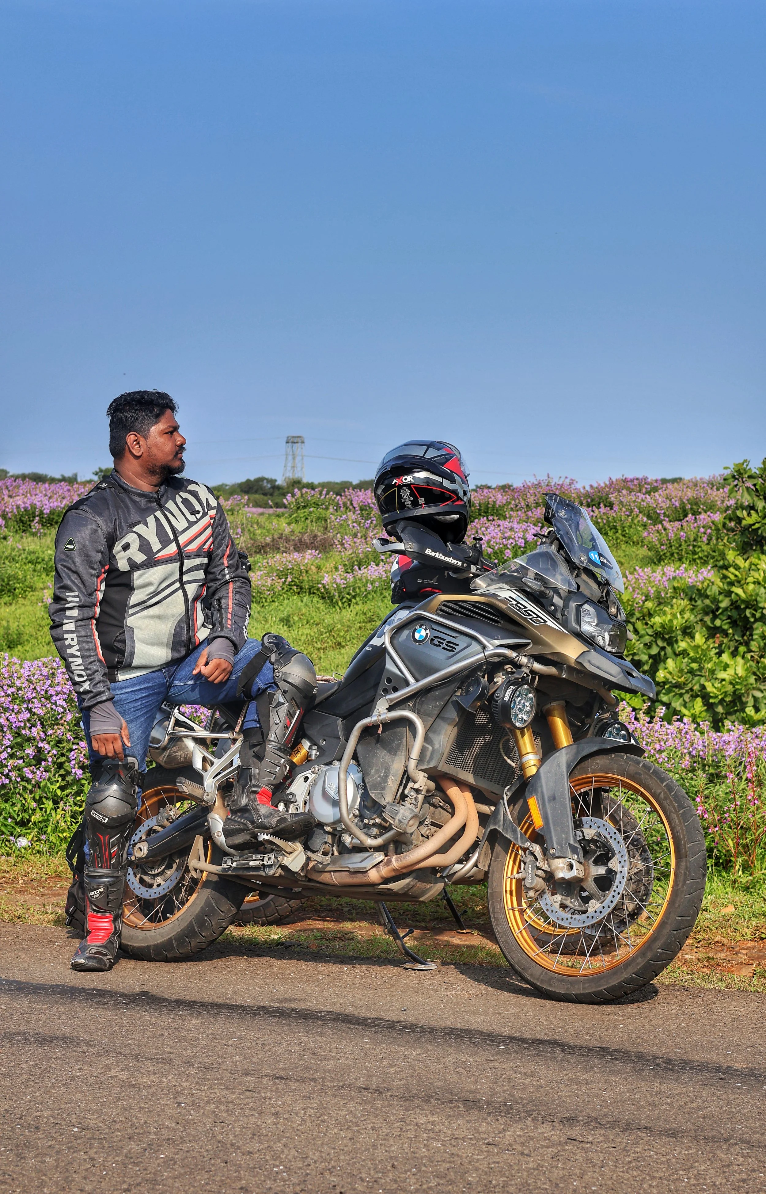 A man dressed in motorcycle gear standing beside a BMW adventure motorcycle on a rural road with flowering bushes and a power line in the background.