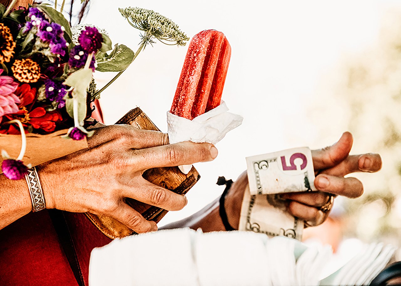 Person holding a bouquet of colorful flowers, a wooden cup, a red ice cream bar, and a five-dollar bill.