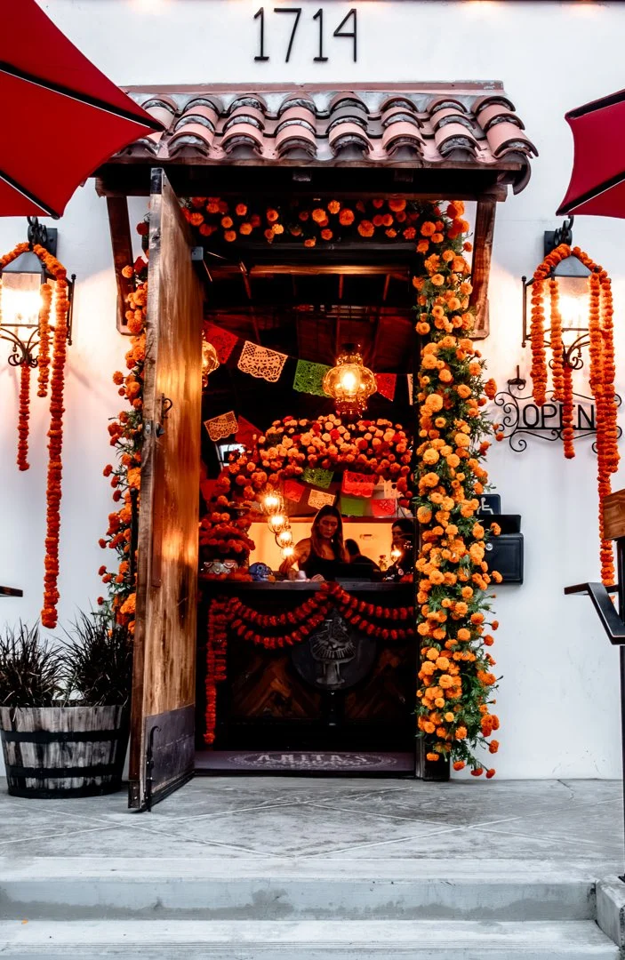 A decorated restaurant entrance with marigold flowers, colorful papel picado banners, and string lights, featuring a woman behind the counter, and a sign that says "OPEN" on the right side.