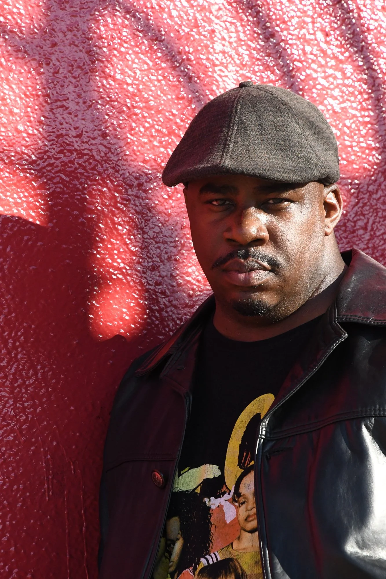 A man wearing a gray flat cap, black leather jacket, and a graphic T-shirt stands in front of a textured red wall with shadows cast on it.