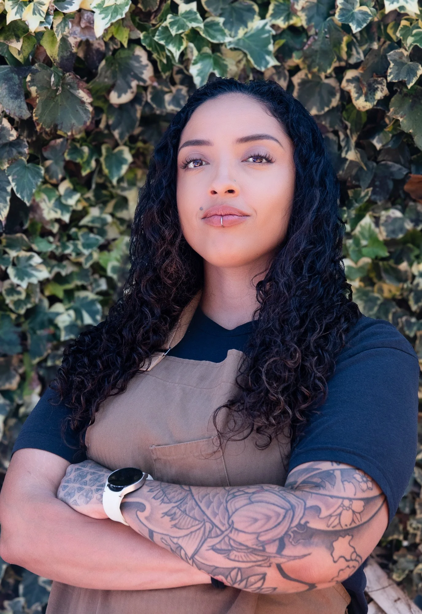 A woman with long, curly dark hair standing outdoors in front of a background of green and variegated ivy leaves. She has a nose and lip piercing, has tattooed arms, and wears a tan apron over a black shirt with a smartwatch on her wrist.