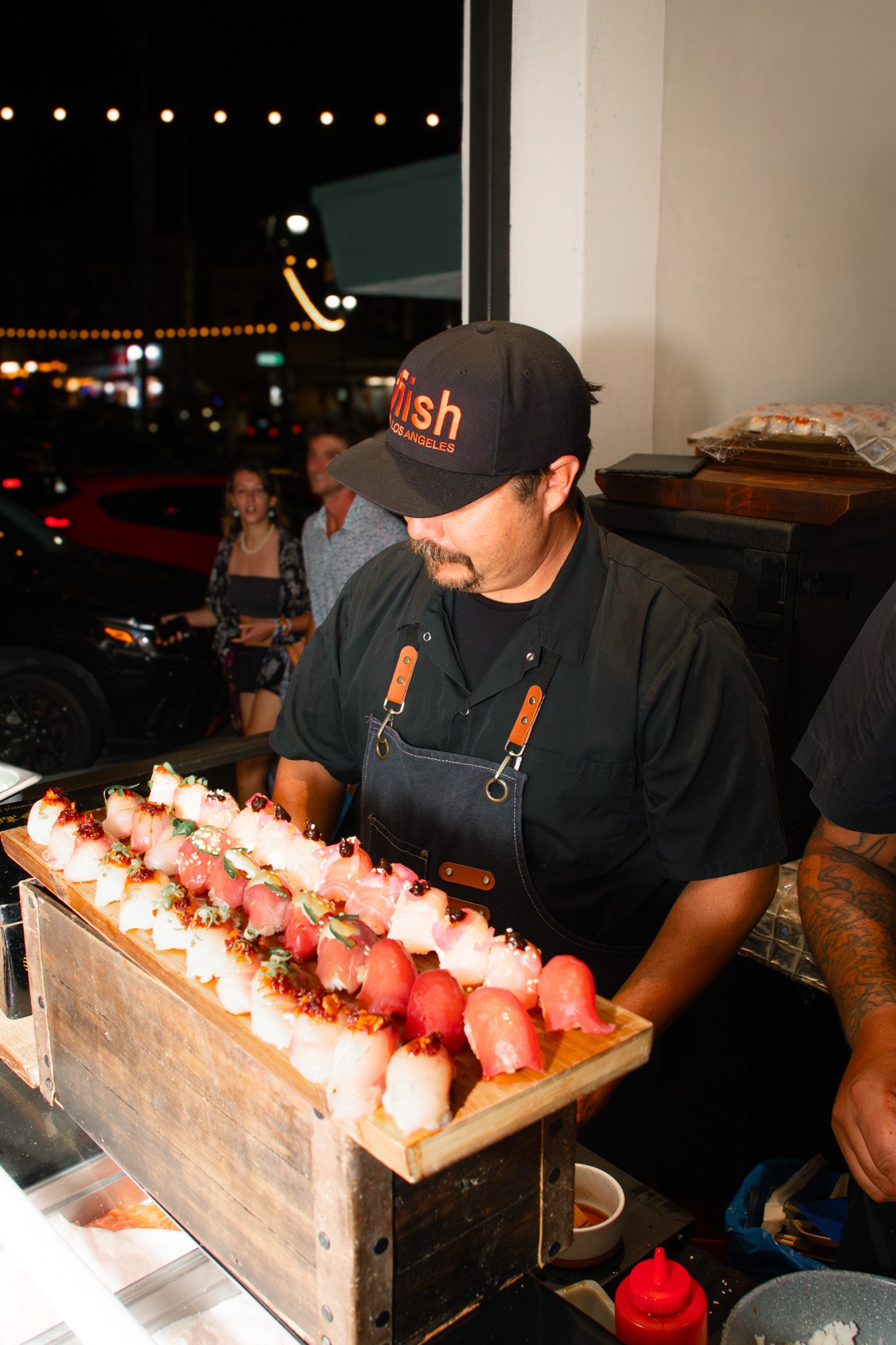 Chef wearing a black cap and apron preparing sushi rolls at an outdoor event at night.