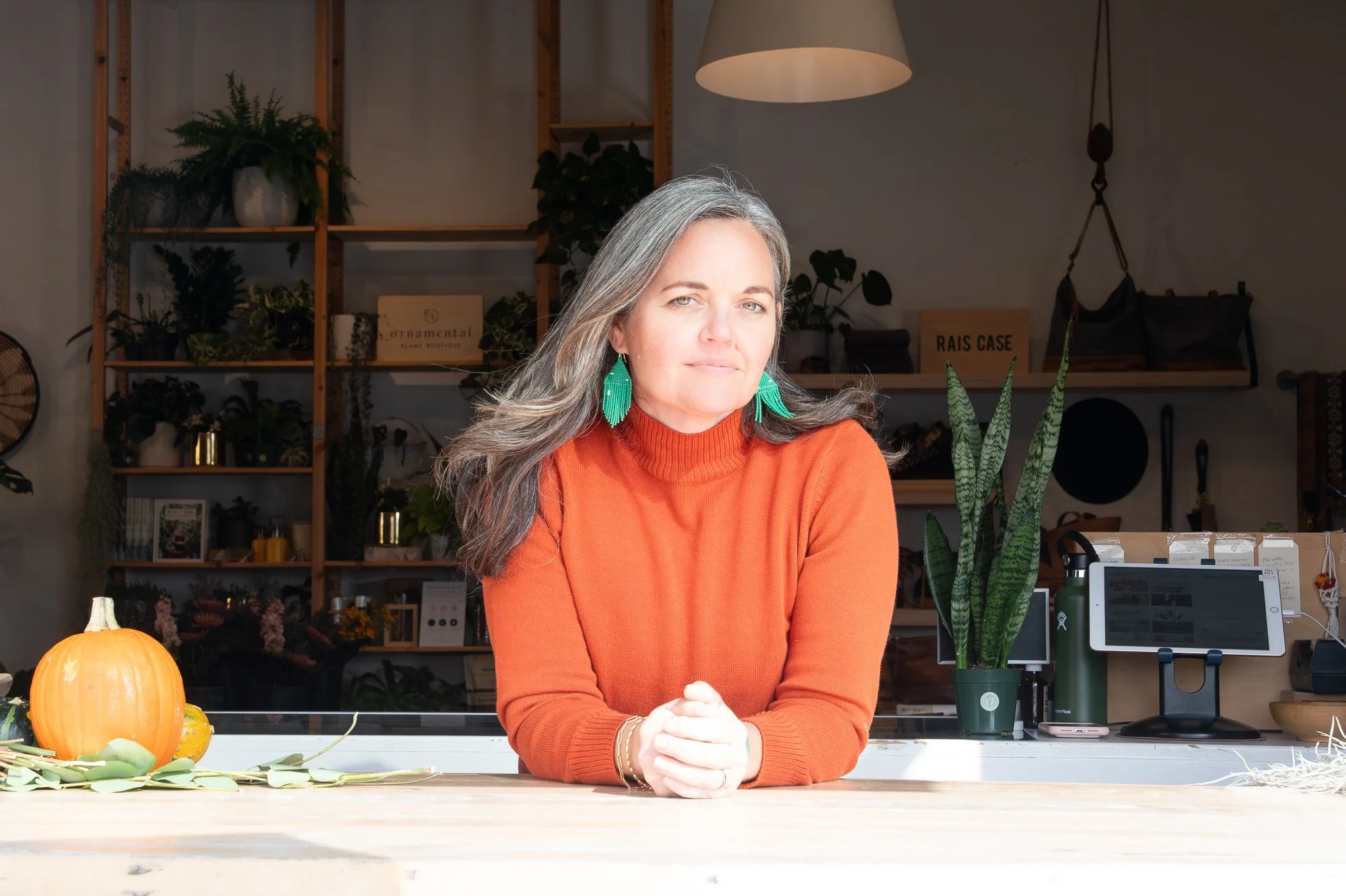 A woman with gray hair, wearing a bright orange sweater and green earrings, is sitting at a light-colored wooden counter with her hands clasped in front of her, in a well-lit space decorated with plants, pumpkins, and various items on shelves behind 