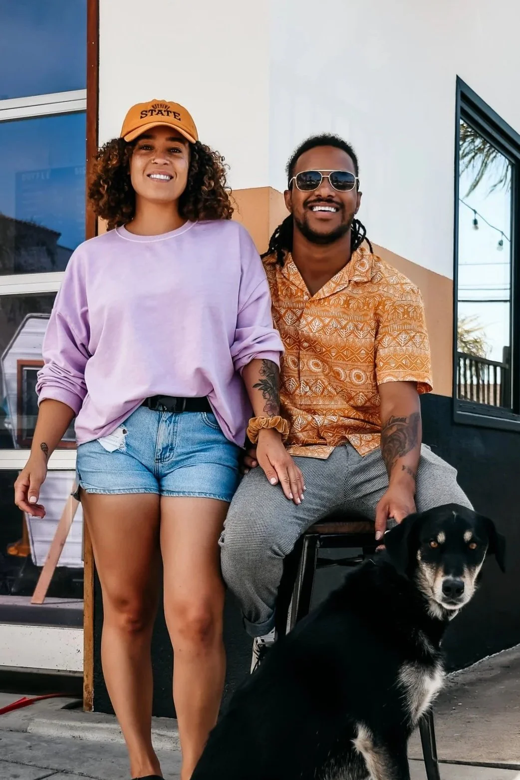 A smiling woman in a lavender oversized sweatshirt, denim shorts, and a mustard cap standing next to a seated man in sunglasses and a patterned shirt with a black and white dog in front of them, outdoors in front of a building with windows.