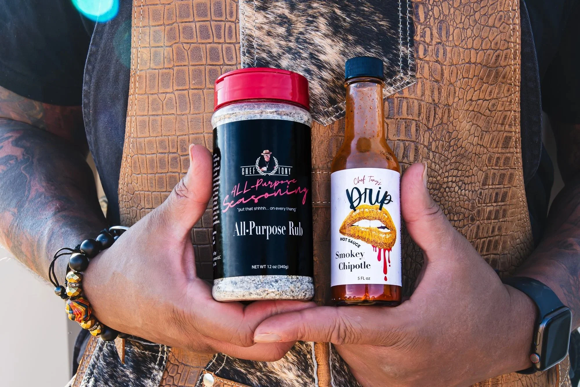 Person holding a container of all-purpose seasoning and a bottle of Smokey Chipotle hot sauce with a leopard print bag and a beaded bracelet.