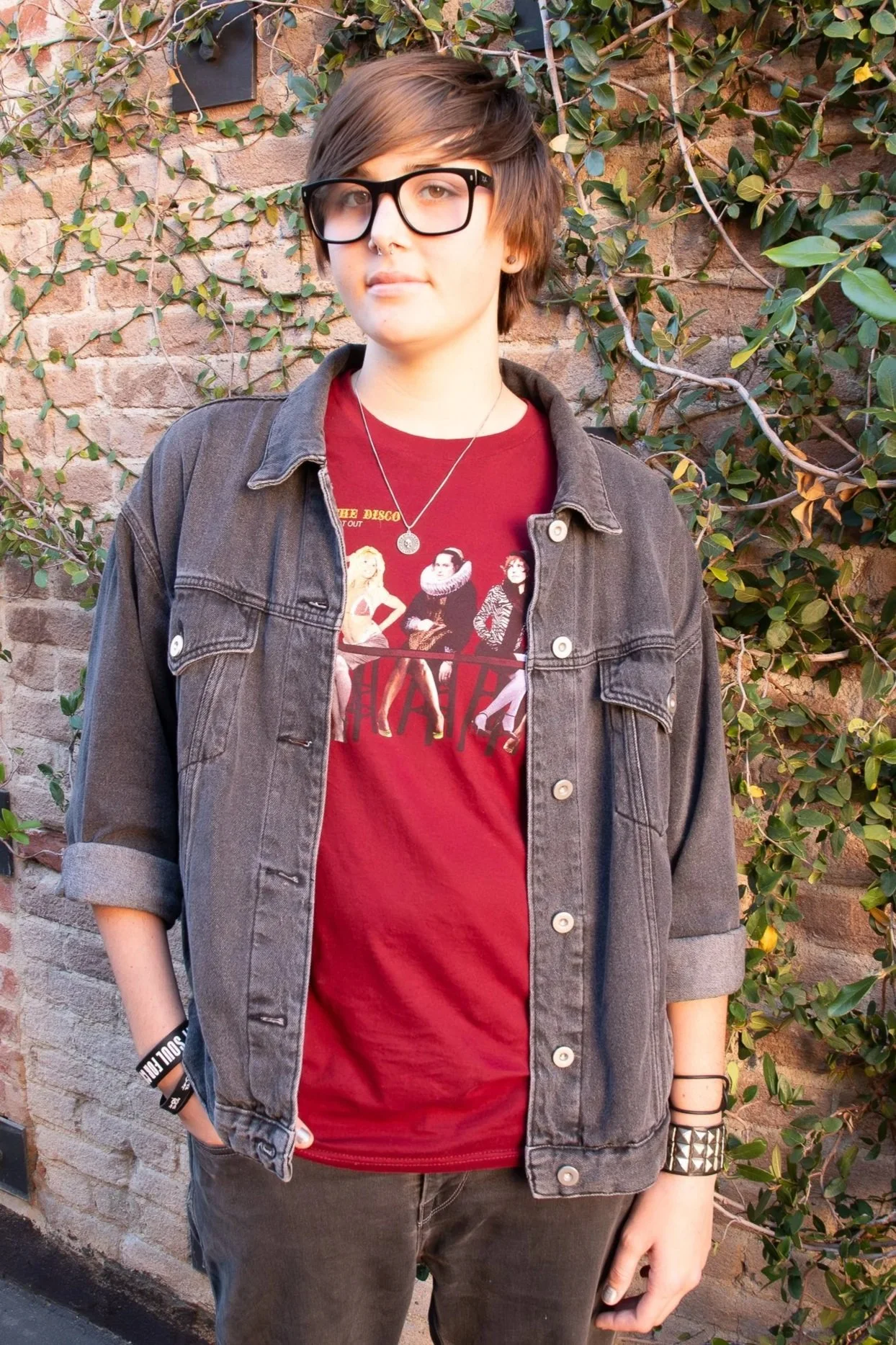 Young person with short brown hair, wearing black glasses, a red shirt featuring a "Hairspray" cast photo, a layered necklace, a gray denim jacket with rolled sleeves, black pants, a black wristband, and a studded bracelet, standing outdoors against 