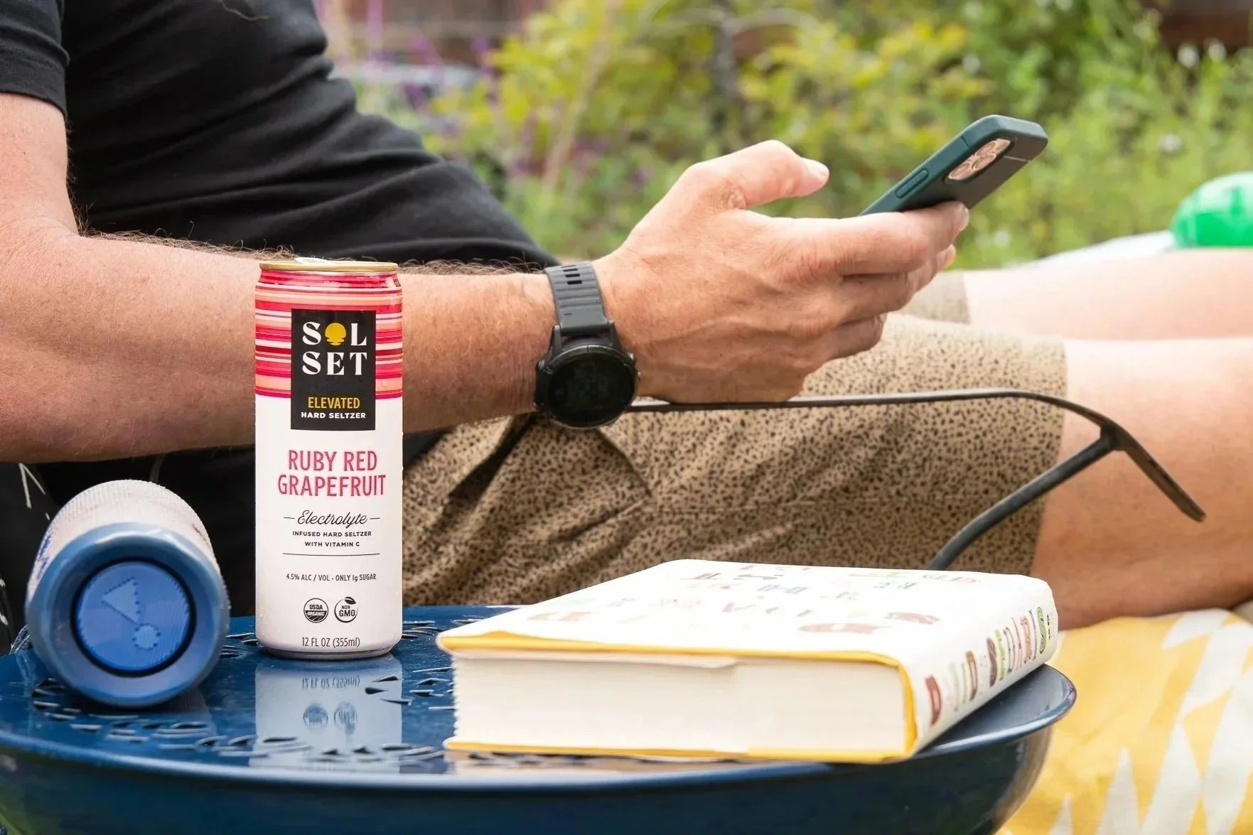 Close-up of a person holding a smartphone, sitting outdoors at a blue table with a can of Solset Ruby Red Grapefruit electrolyte seltzer, a bottle lying on its side, and a closed book.
