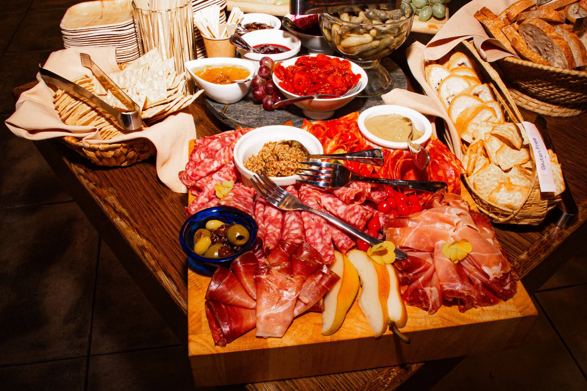 A wooden table set with a variety of breakfast foods including sliced bread, cured meats, cheeses, fruit spreads, olives, cherry tomatoes, and several bowls of jam and honey, with baskets of bread and crackers.