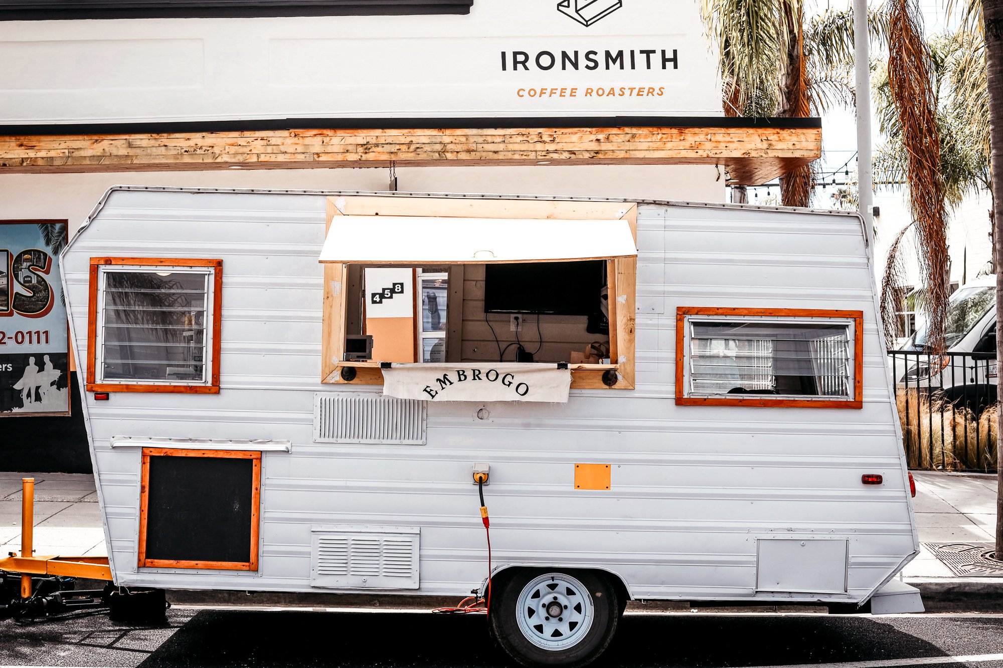 White food truck with orange trim featuring a service window, a chalkboard menu, and a banner that reads 'EMBARGO', parked outside a coffee shop named 'IRONSINTH COFFEE ROASTERS'.