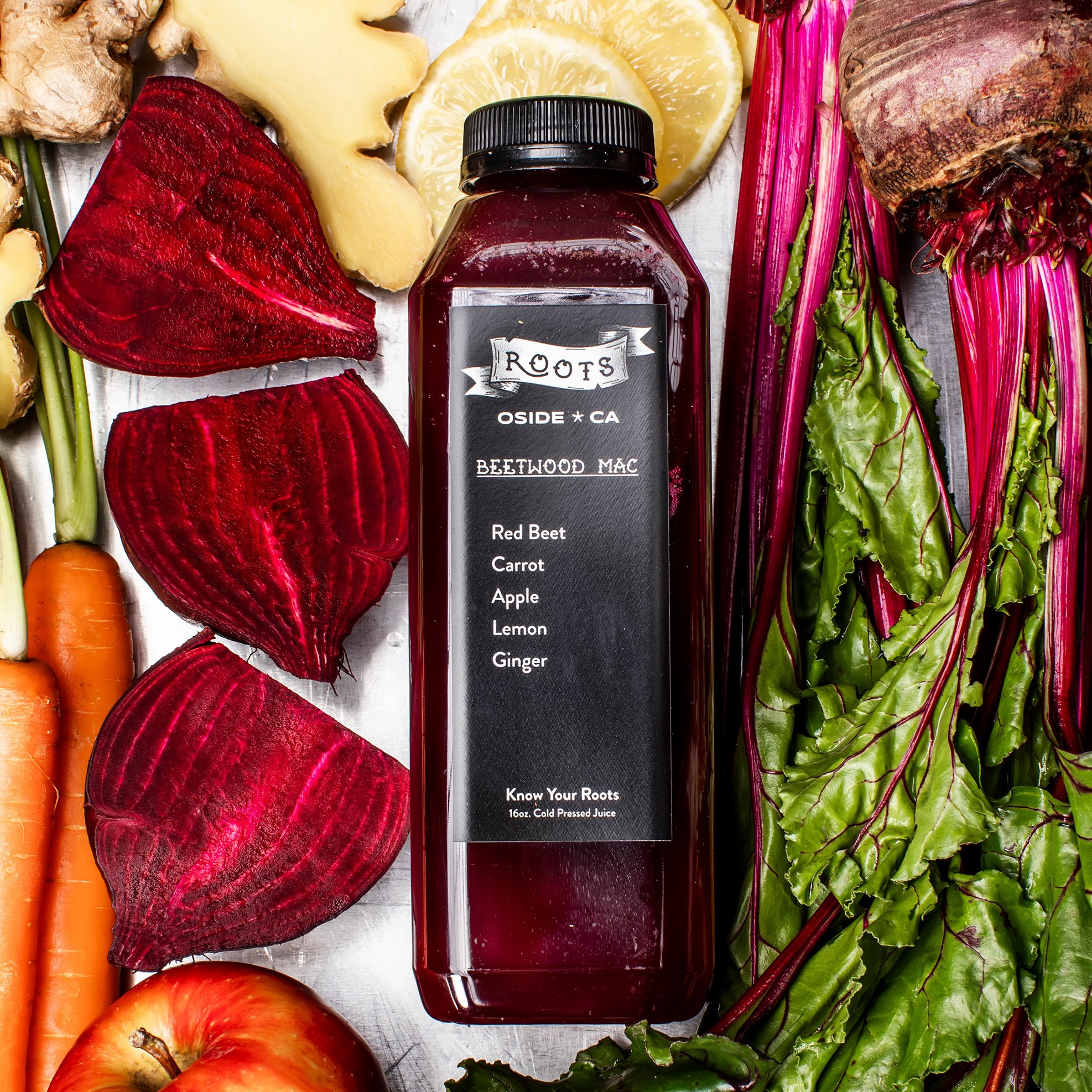 Bottle of Roots beetroot juice surrounded by fresh vegetables and fruits including beets, carrots, apples, lemons, ginger, and leafy greens.