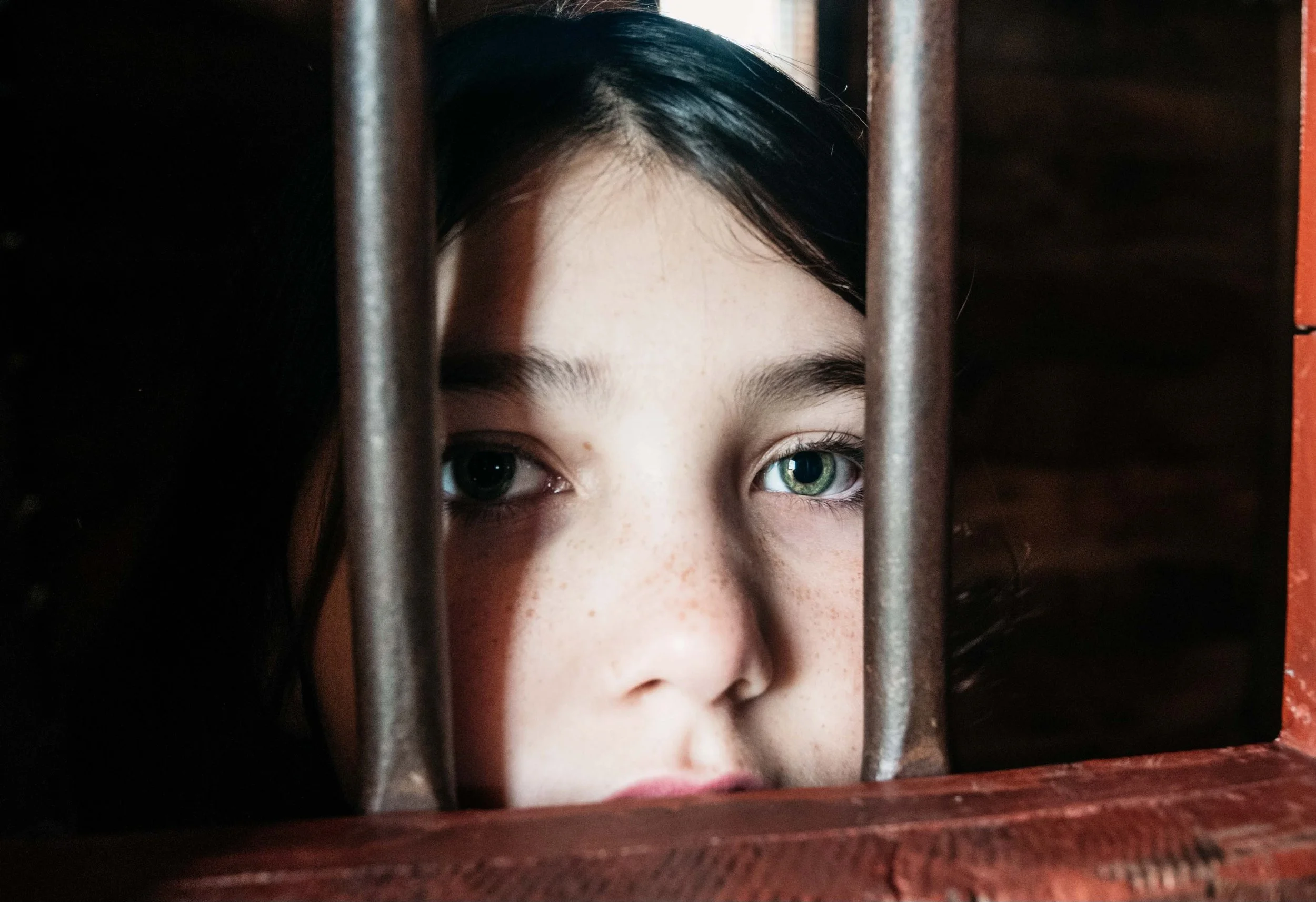 Close-up of a young girl with blue eyes looking through metal bars of a cage, with a wooden structure on the side.