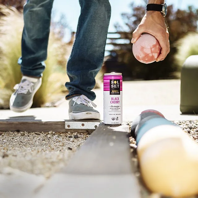 Person standing outdoors on a wooden plank while holding a noose with a peach in hand, with a can of Solset Black Cherry soda and a cylindrical object on the ground.