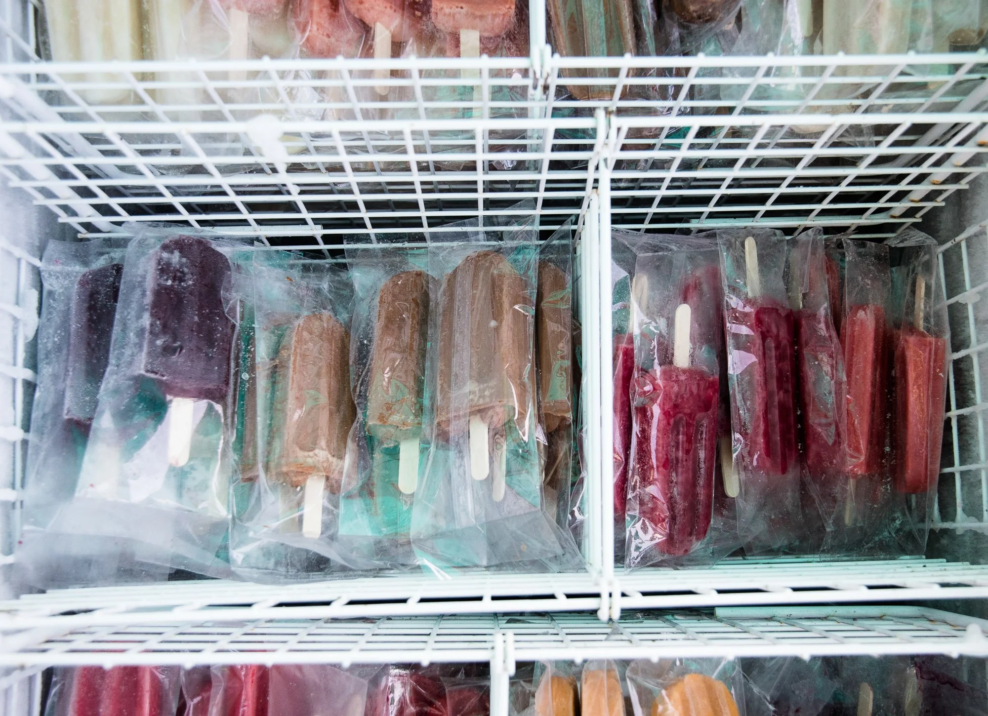 A top-down view of a wire freezer basket filled with individually wrapped popsicles in various shades of red, purple, brown, and orange.