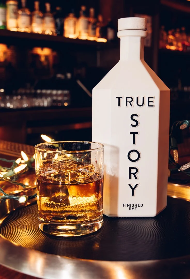 A glass of finished rye whiskey with ice and a white bottle labeled "True Story" on a table in a dimly lit bar.