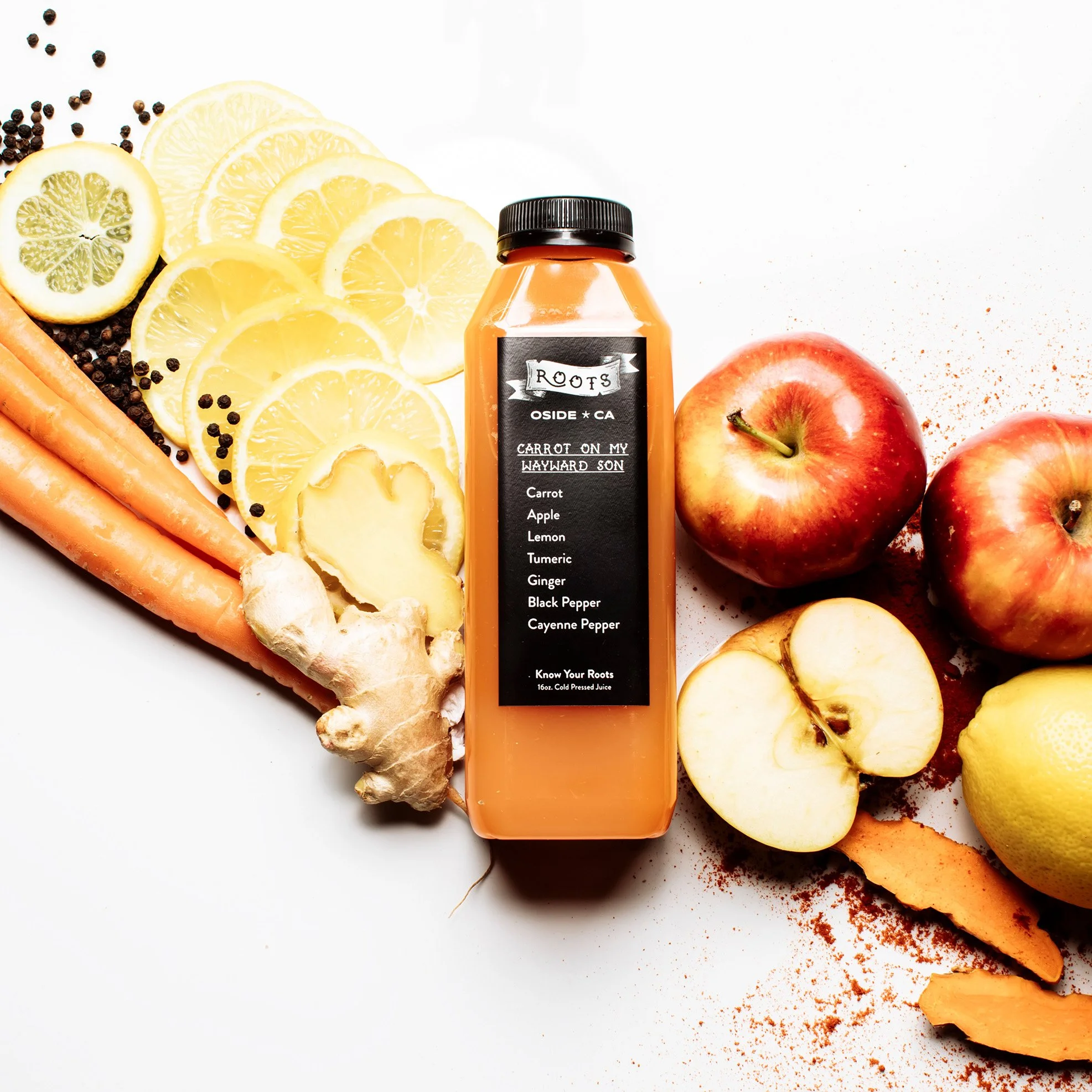 Juice bottle surrounded by sliced lemons, oranges, apples, carrots, ginger, and pepper on a white surface.