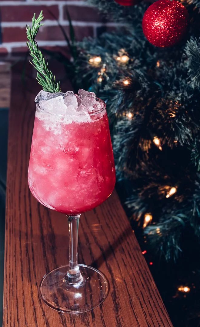 A pink Christmas cocktail with crushed ice and a sprig of rosemary in a stemmed glass on a wooden surface, with a decorated Christmas tree and brick wall in the background.