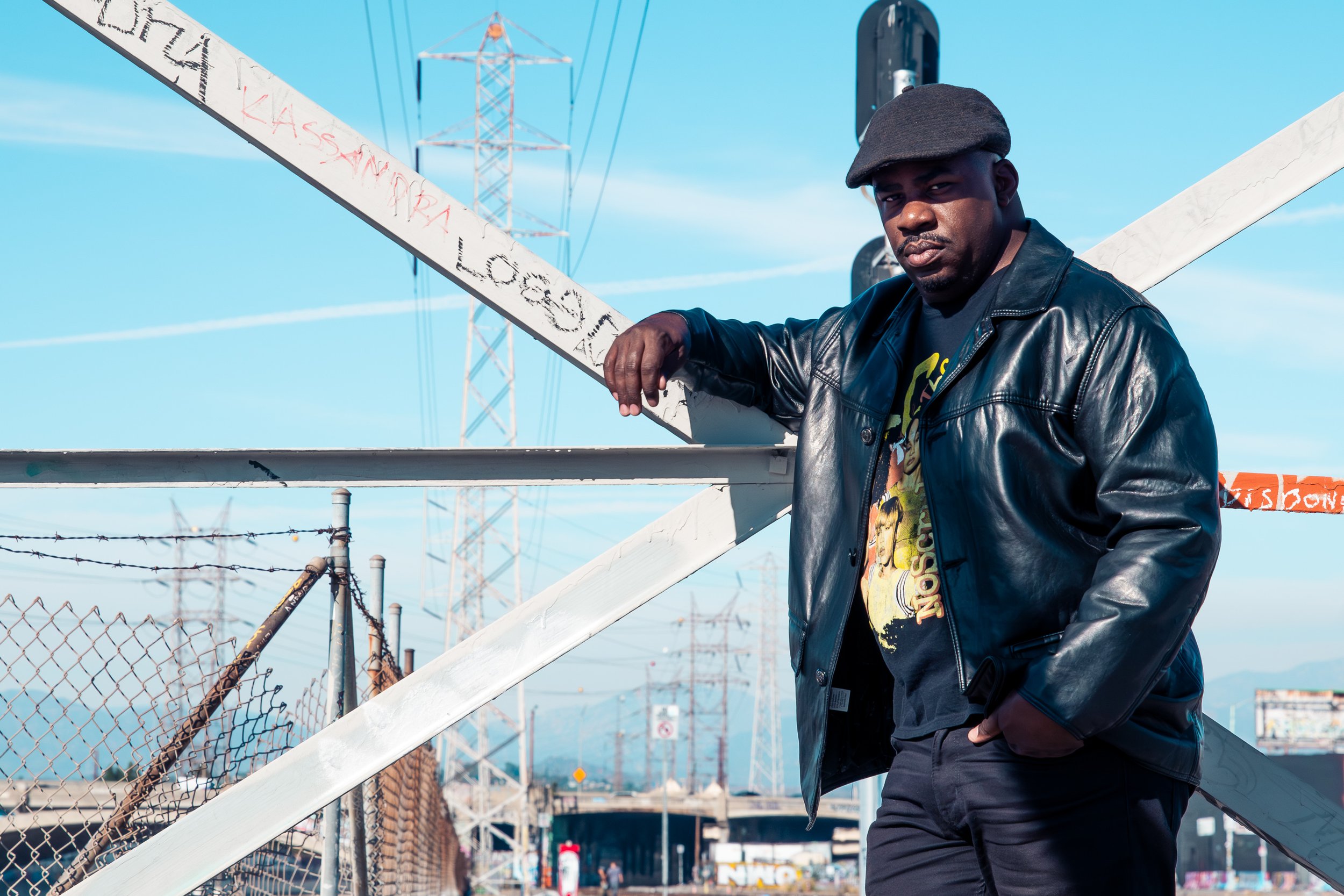A man in a black leather jacket, graphic t-shirt, and dark cap standing near a chain-link fence and leaning on a white metal structure in an outdoor urban setting, with power lines and clear blue sky in the background.