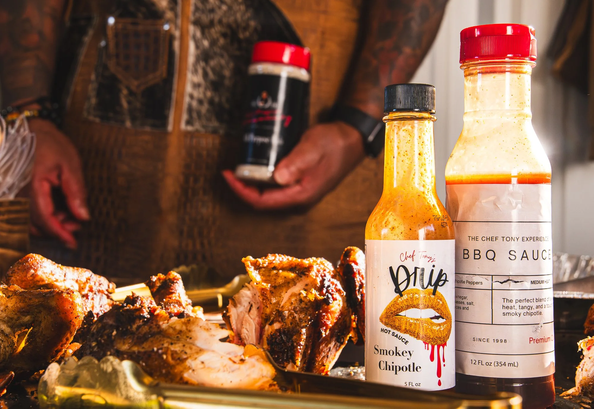 Barbecue chicken with sauces including smoky chipotle hot sauce and BBQ sauce on a platter, with a person in the background holding a container of seasoning.