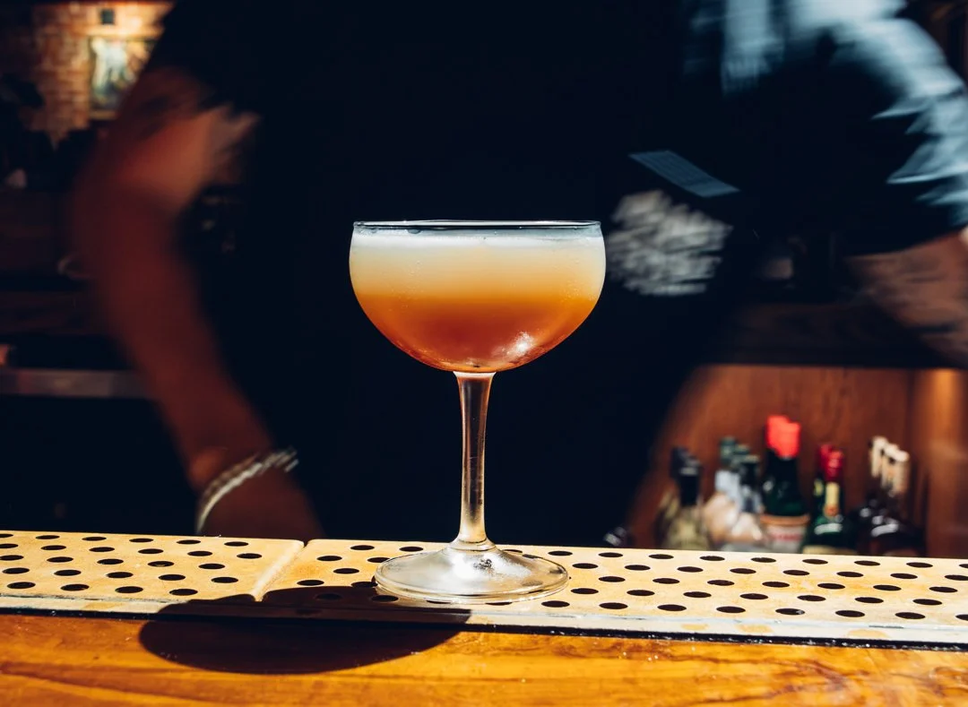 A cocktail in a coupe glass with a layered orange and red hue, placed on a bar counter.