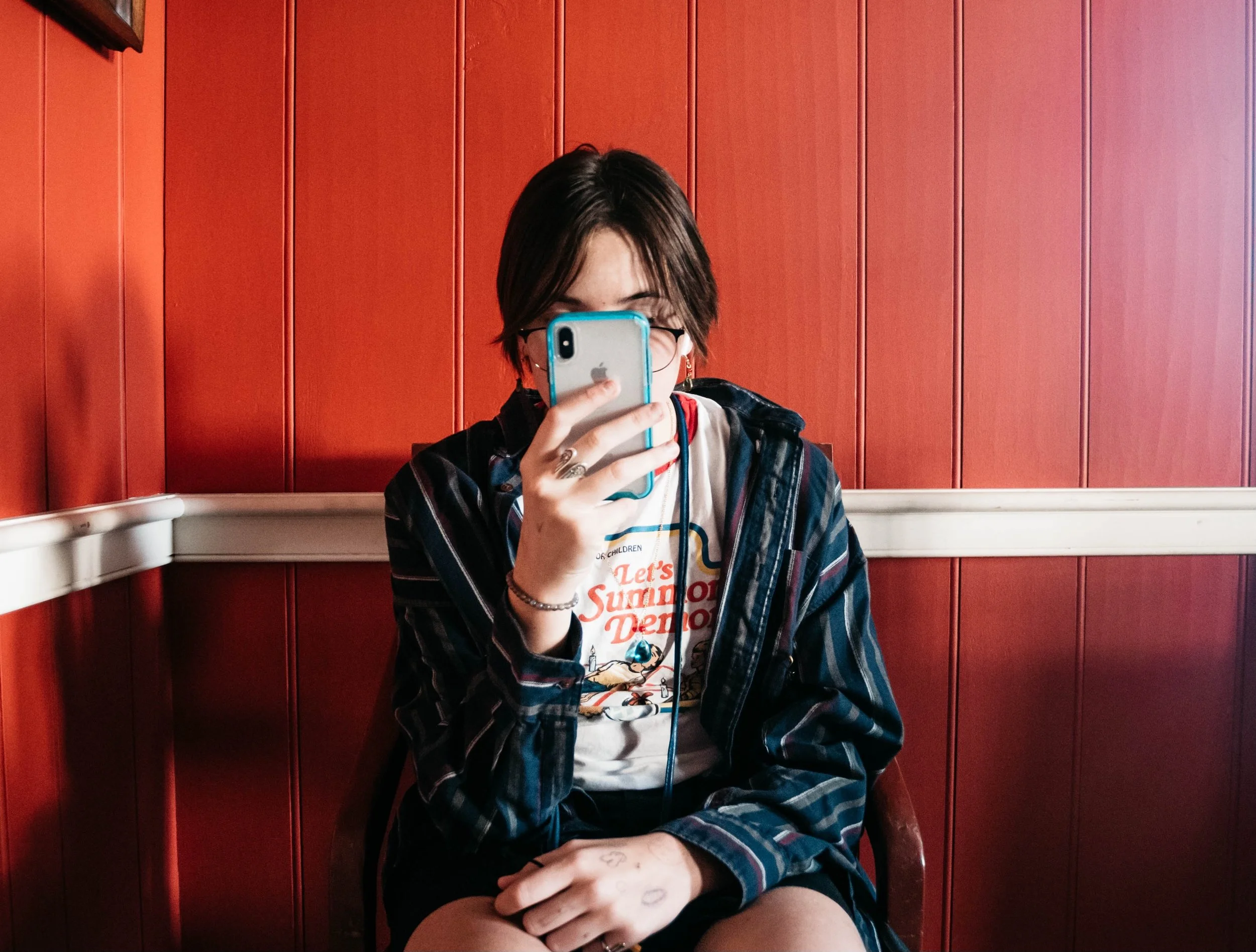 A person with short dark hair wearing glasses, a graphic T-shirt, and a striped jacket, taking a mirror selfie with a smartphone inside a room with red wood-paneled walls.