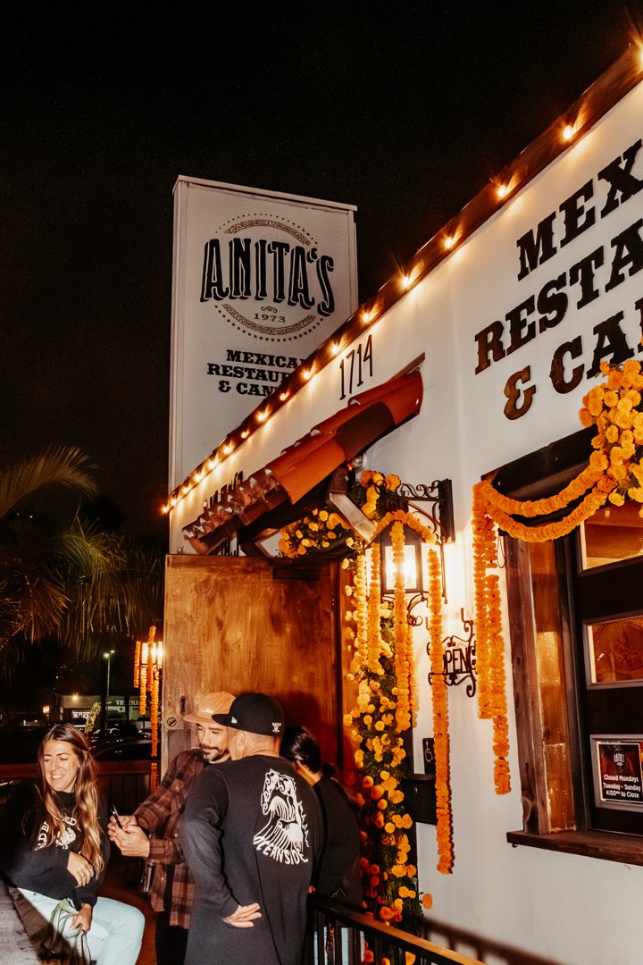 People gathered outside Anita's Mexican restaurant decorated with marigold flowers for a celebration, at night.
