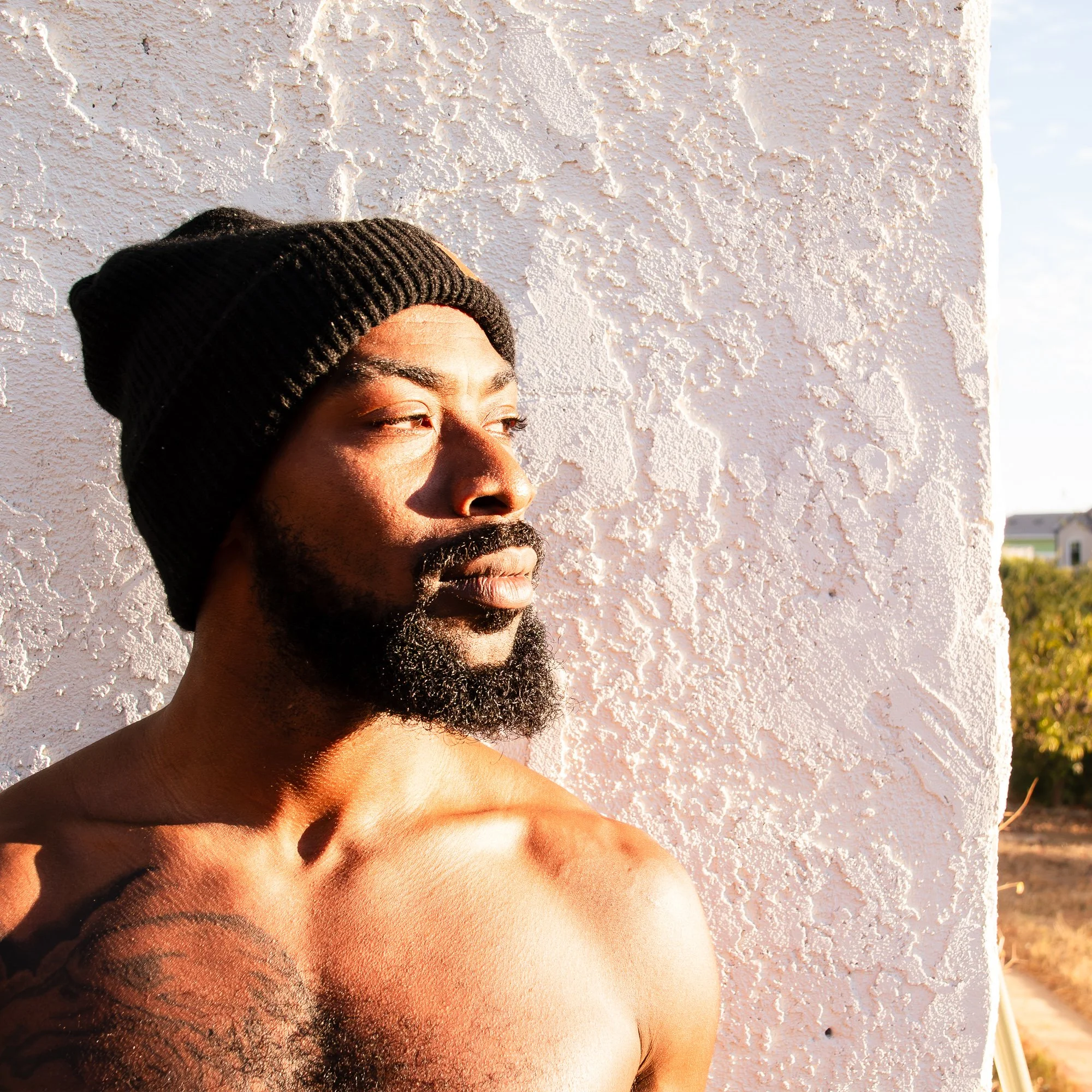 A shirtless man with a beard and a black knit cap leaning against a textured white wall, gazing into the distance during sunset.