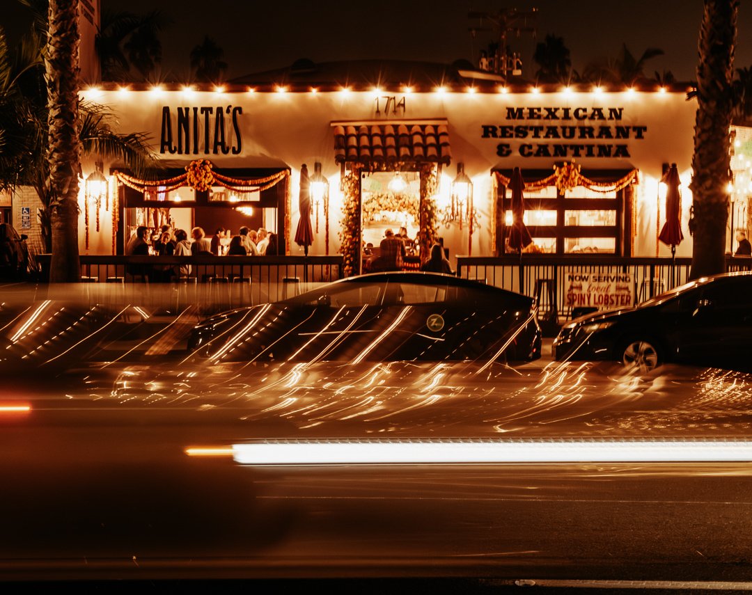 Nighttime view of Anita's Mexican Restaurant & Cantina with people dining inside, palm trees on either side, and cars parked in front. Light streaks from passing vehicles are visible in the foreground.