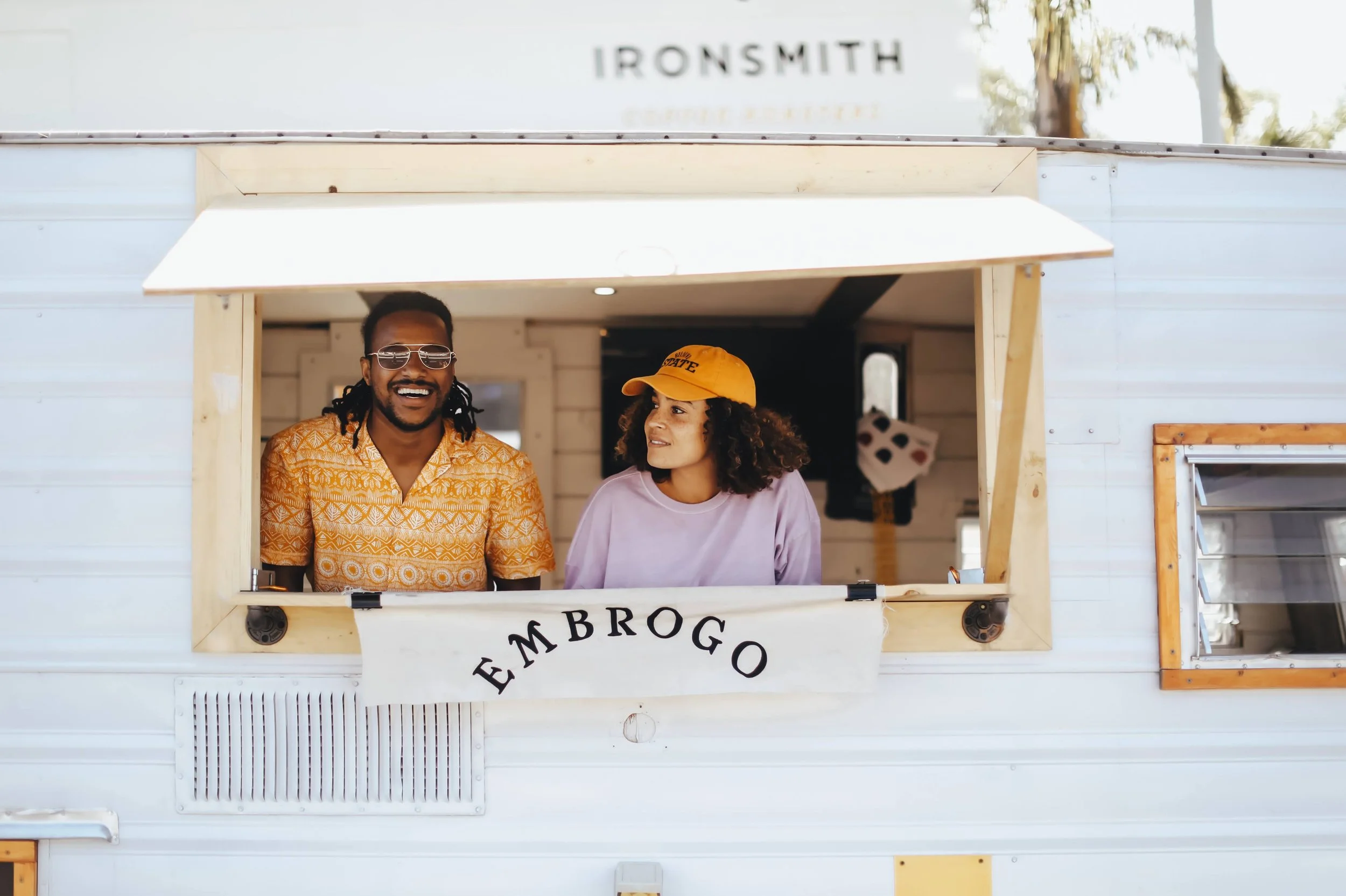 Two people are inside a food truck named EMbrogO, smiling and talking to each other. The man has dreadlocks, wears sunglasses, and a patterned shirt, while the woman has curly hair, a yellow cap, and a pastel purple shirt.