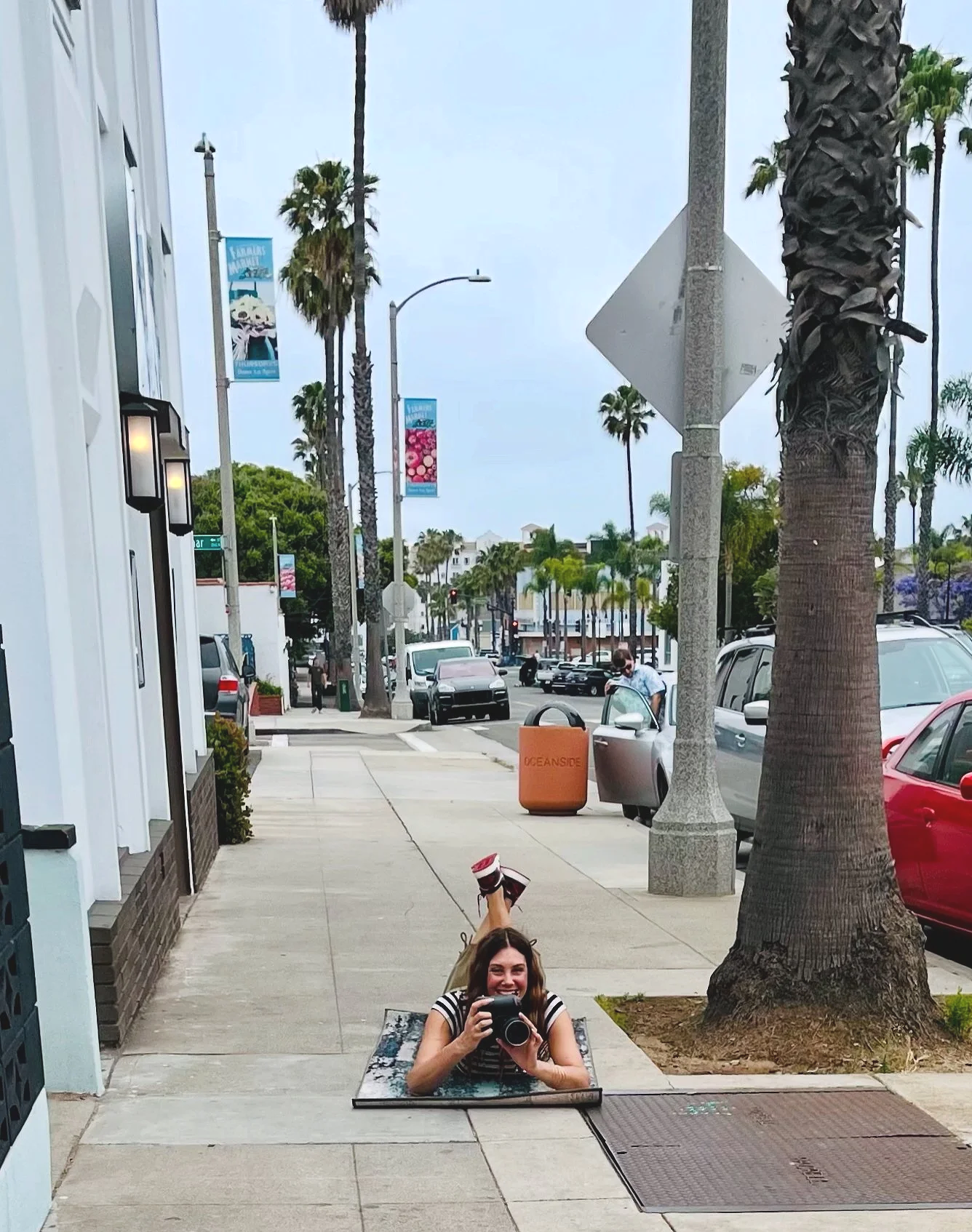 A woman lying on her stomach on the sidewalk with her head and arms through a rectangular opening, smiling and taking a photograph of herself with a camera, with her legs raised behind her. She is wearing a striped shirt, and the scene is set in a city street lined with palm trees and parked cars.