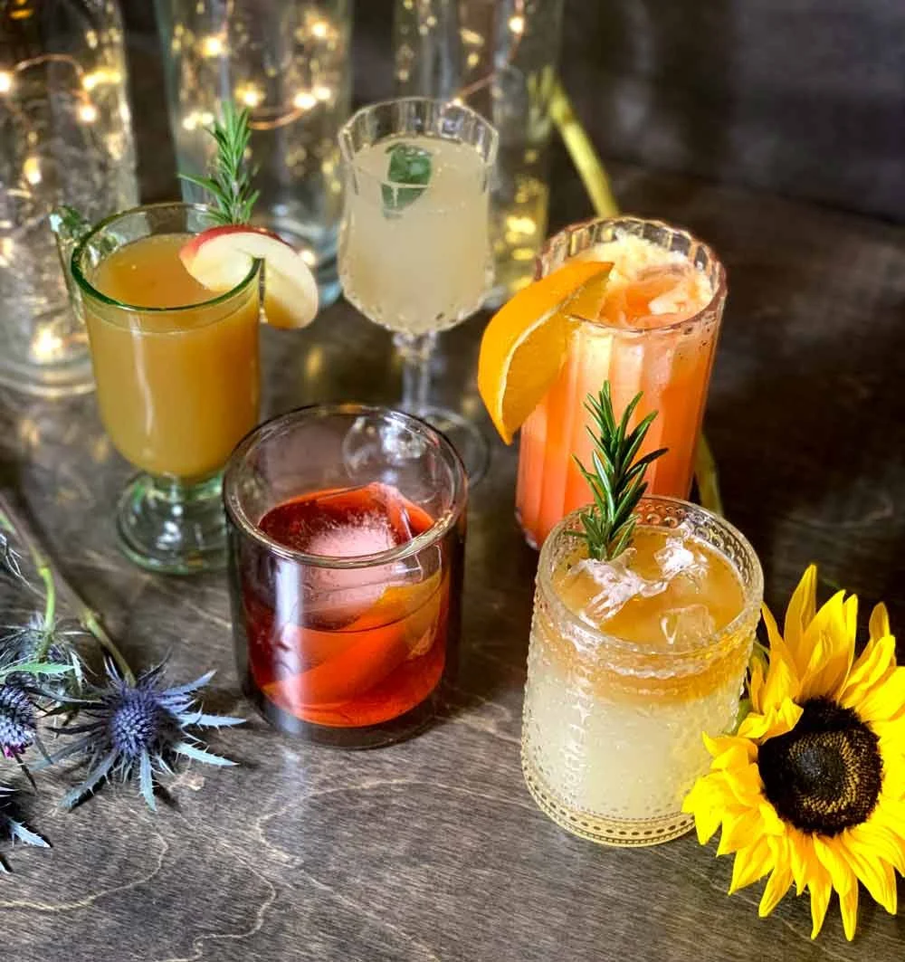 Six colorful cocktails with garnishes including fruit slices and herbs arranged on a wooden table, decorated with sunflowers and purple thistle flowers.