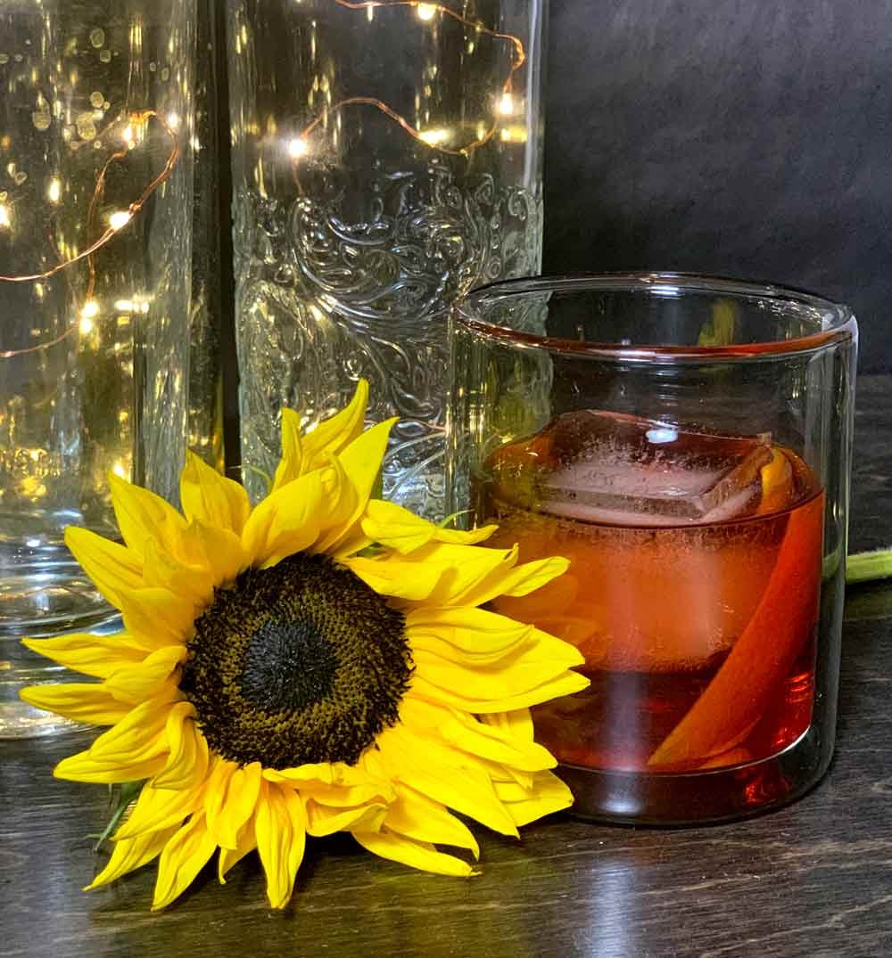 A yellow sunflower with dark center lying on a dark wooden surface next to a glass of iced amber-colored tea with ice cubes and an orange slice, with glass jars in the background decorated with string lights.