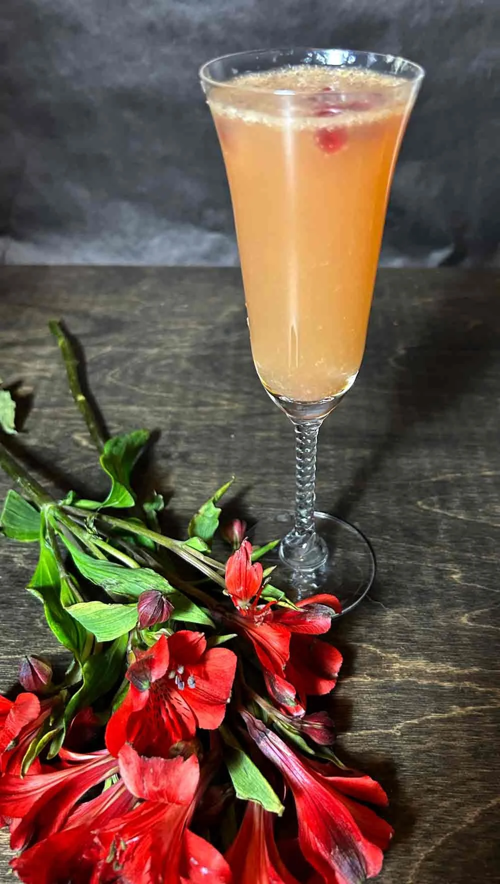 A tall, elegant champagne flute filled with a pinkish-orange cocktail, garnished with red pomegranate seeds, set on a dark wooden surface next to a small bouquet of red flowers and green foliage.
