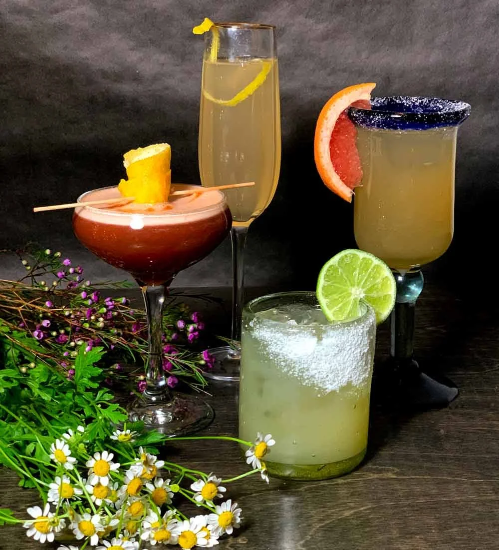 Four different cocktails with garnishes, surrounded by flowers on a dark surface.