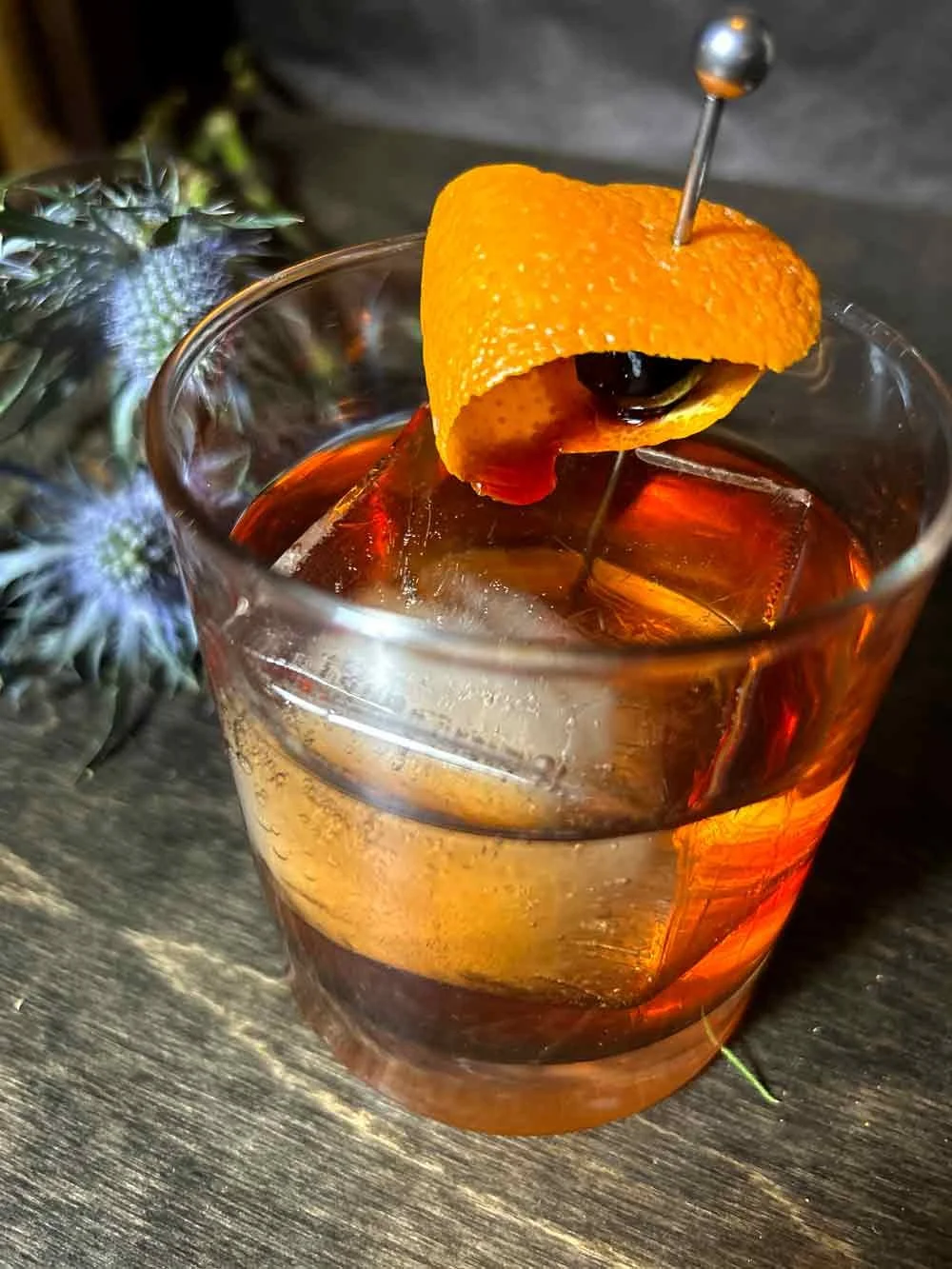 A glass of cocktail with ice, garnished with an orange peel and a black cherry, secured with a cocktail pick, with a pineapple plant on the side.