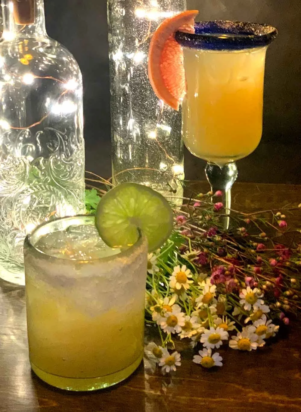 Two cocktails with citrus slices and flowers, decorative bottles with fairy lights in the background.