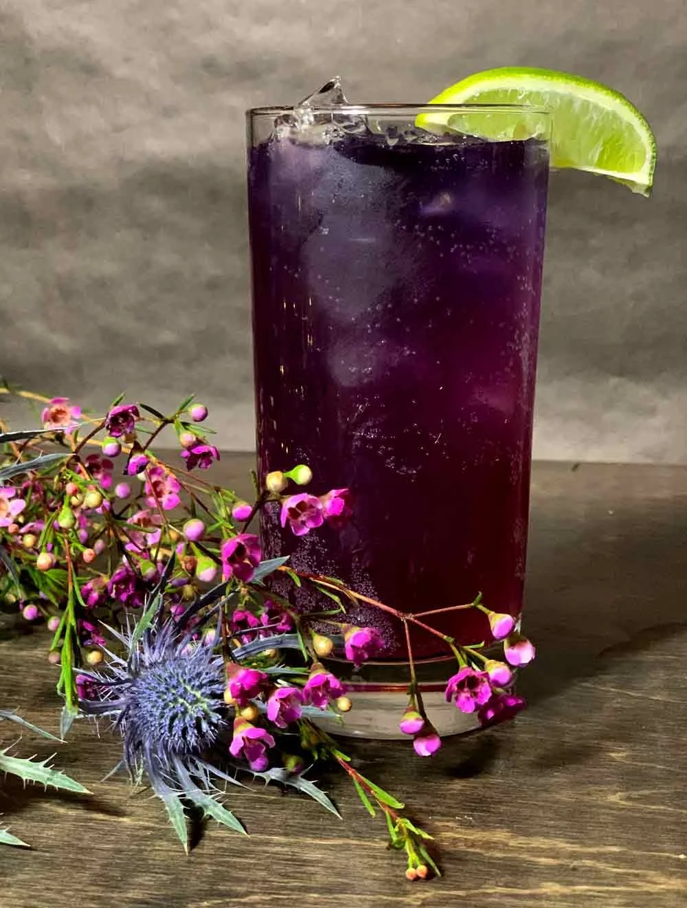 A tall glass of purple soda with ice, garnished with a lime wedge, resting on a wooden surface next to purple and green flowers and spiky foliage.