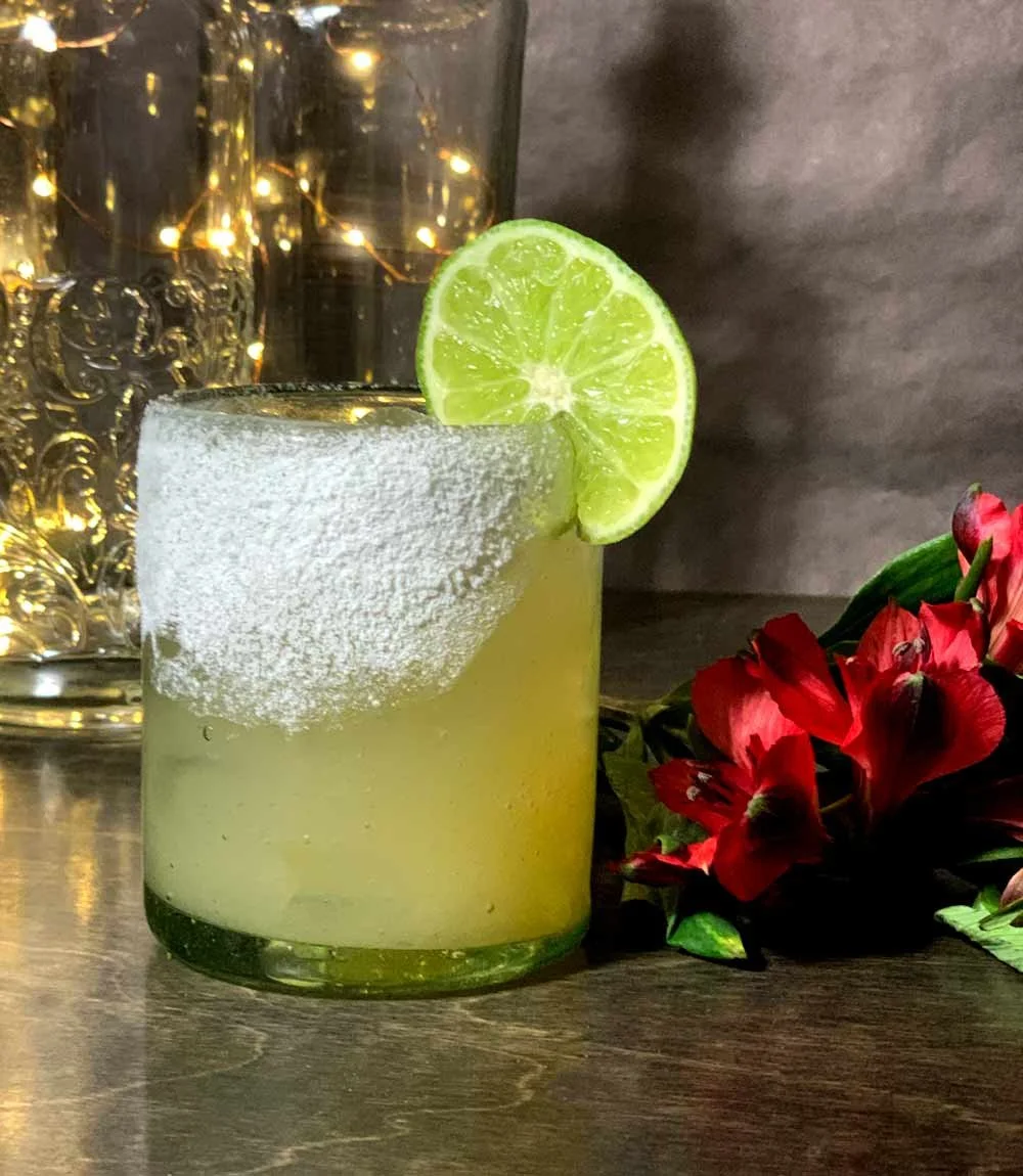 A cocktail in a glass with a salted rim and a lime wedge garnish, placed on a wooden surface with red flowers beside it.