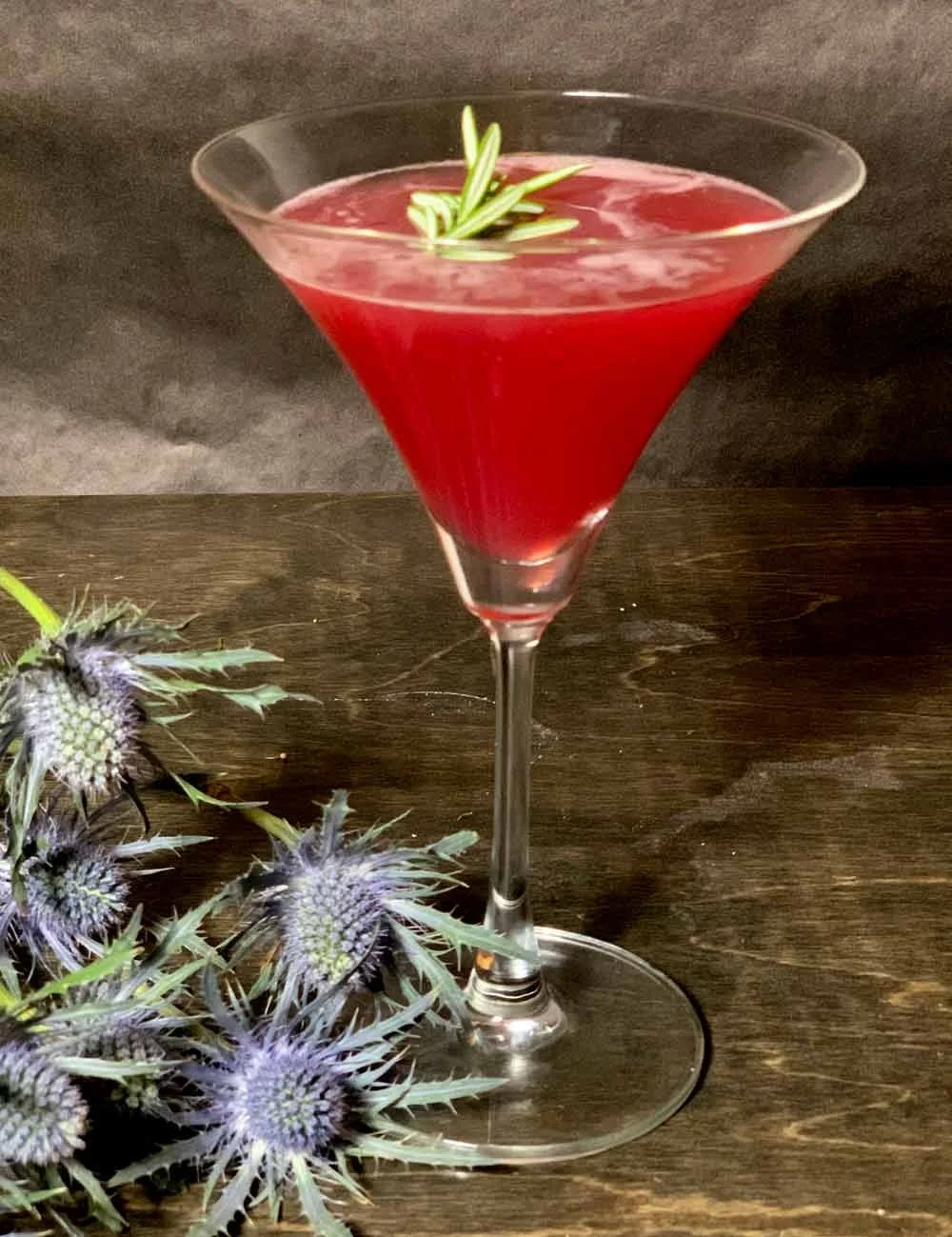 A red cocktail garnished with a sprig of rosemary in a martini glass on a wooden surface, with purple thistle flowers nearby.