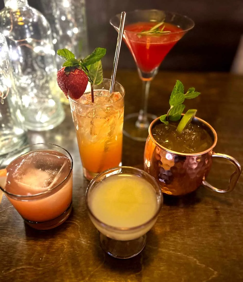 Assorted colorful cocktails on a wooden bar table, garnished with fruit, mint, and decorated glasses.
