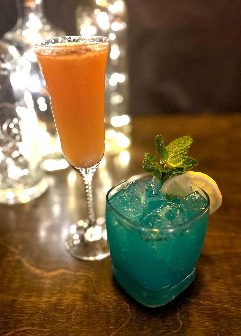 A blue cocktail with ice, lemon slice, and mint leaves garnish in a glass, and an orange-colored drink in a tall champagne flute on a wooden table, with blurred lights in the background.