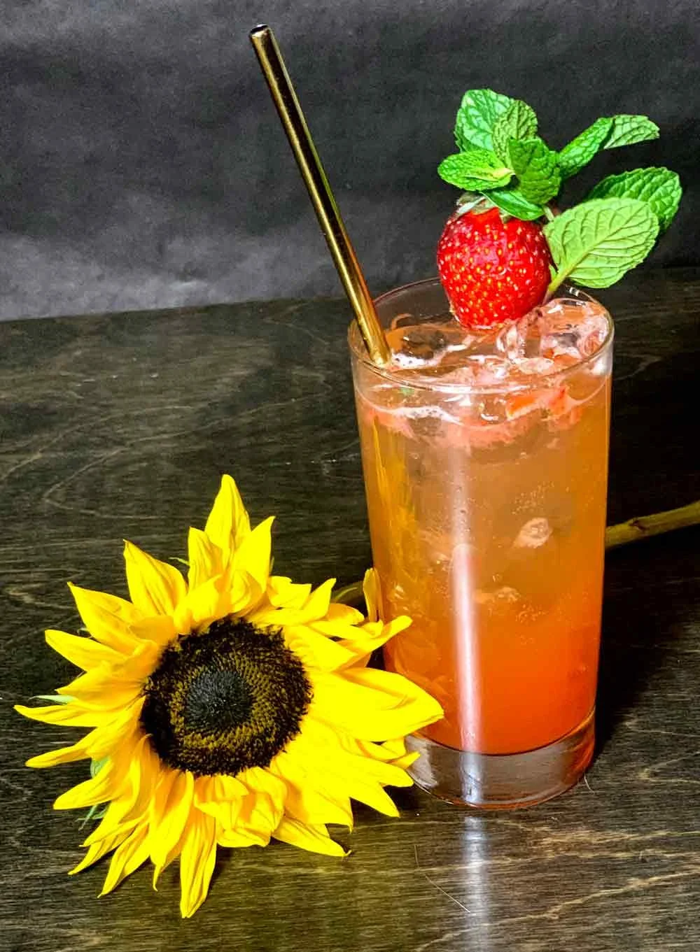 A tall glass of pink strawberry cocktail with ice, garnished with a strawberry, mint leaves, and a gold straw, placed on a dark wooden surface next to a sunflower.