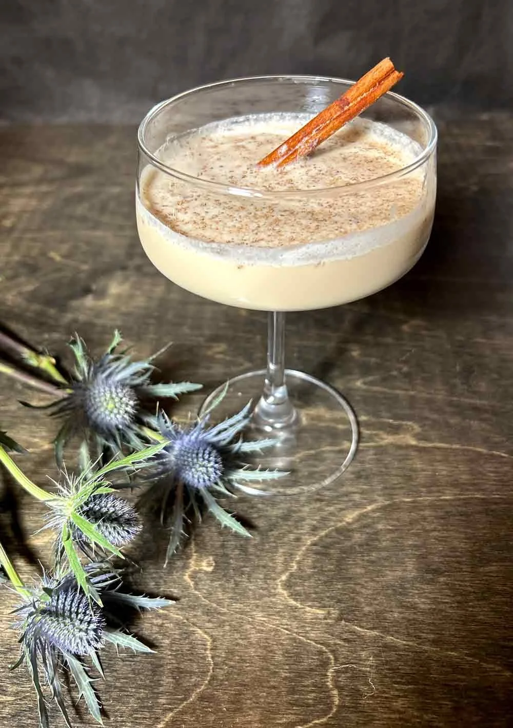 A cocktail in an elegant coupe glass garnished with a cinnamon stick, positioned on a wooden surface, with blue thistle flowers nearby.